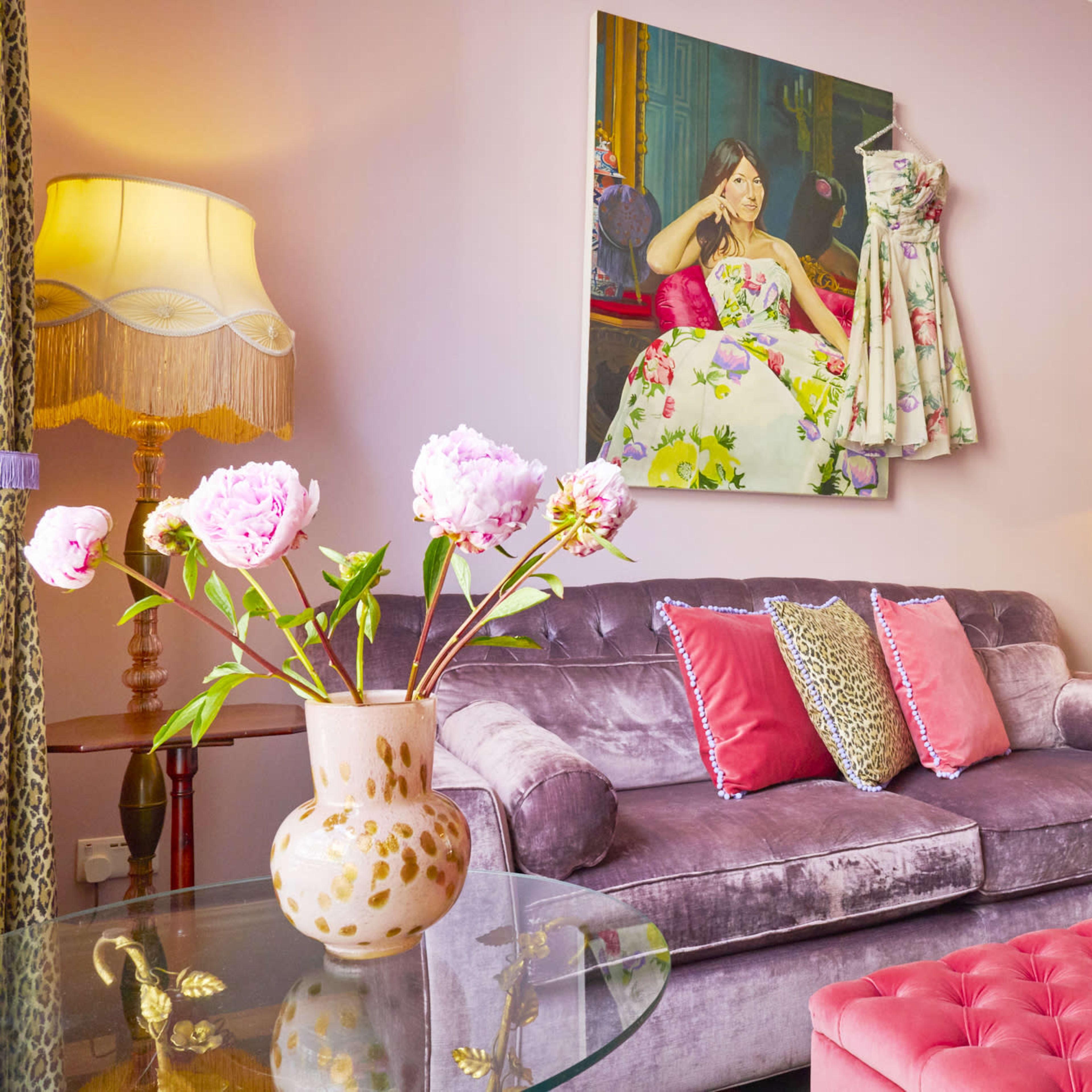 A plush purple sofa with vibrant pillows is placed in a brightly colored room featuring floral artwork and a glass coffee table with a vase of pink peonies.