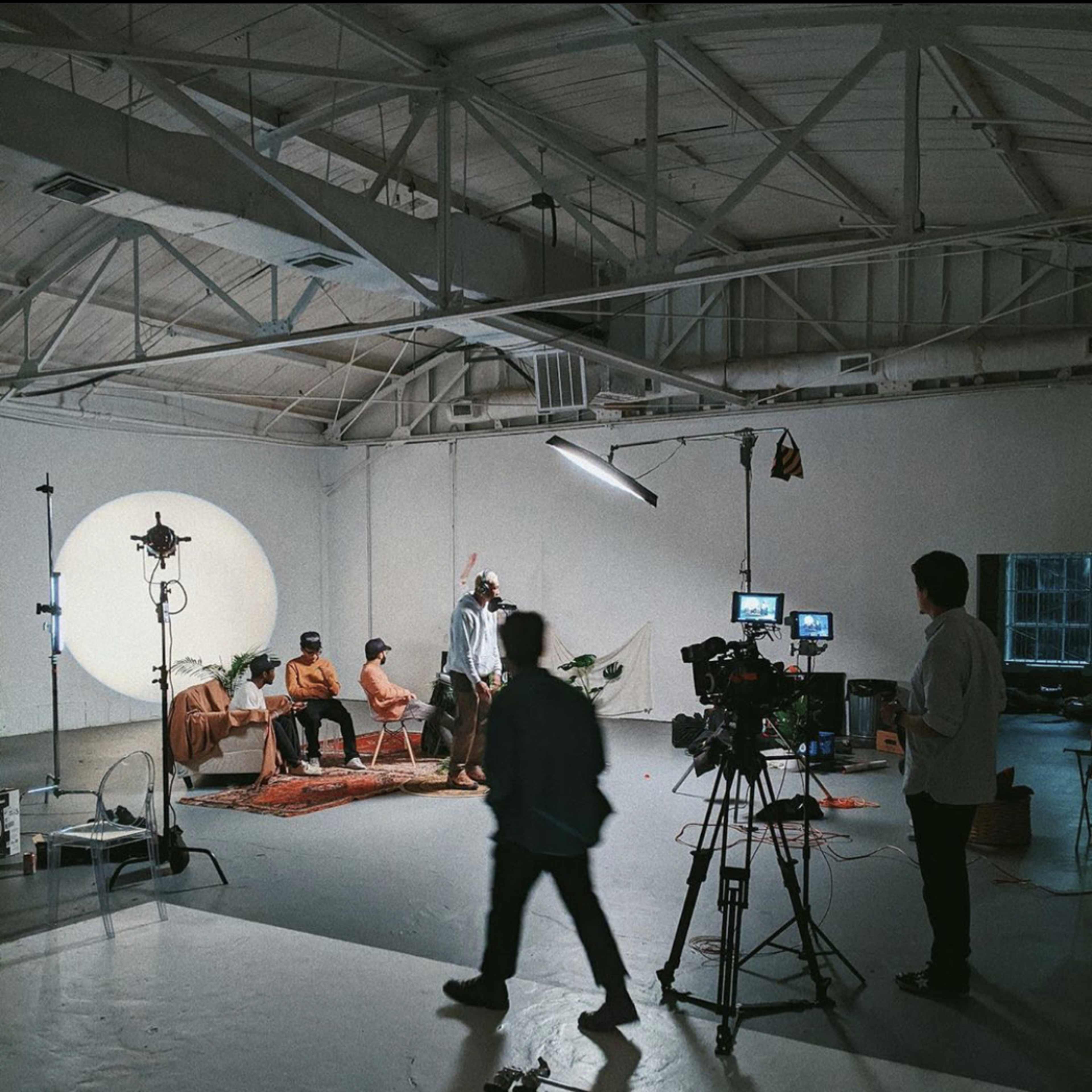 A film crew captures a scene with a group of four individuals sitting on chairs in a minimalist studio with a circular backdrop and various cameras set up.