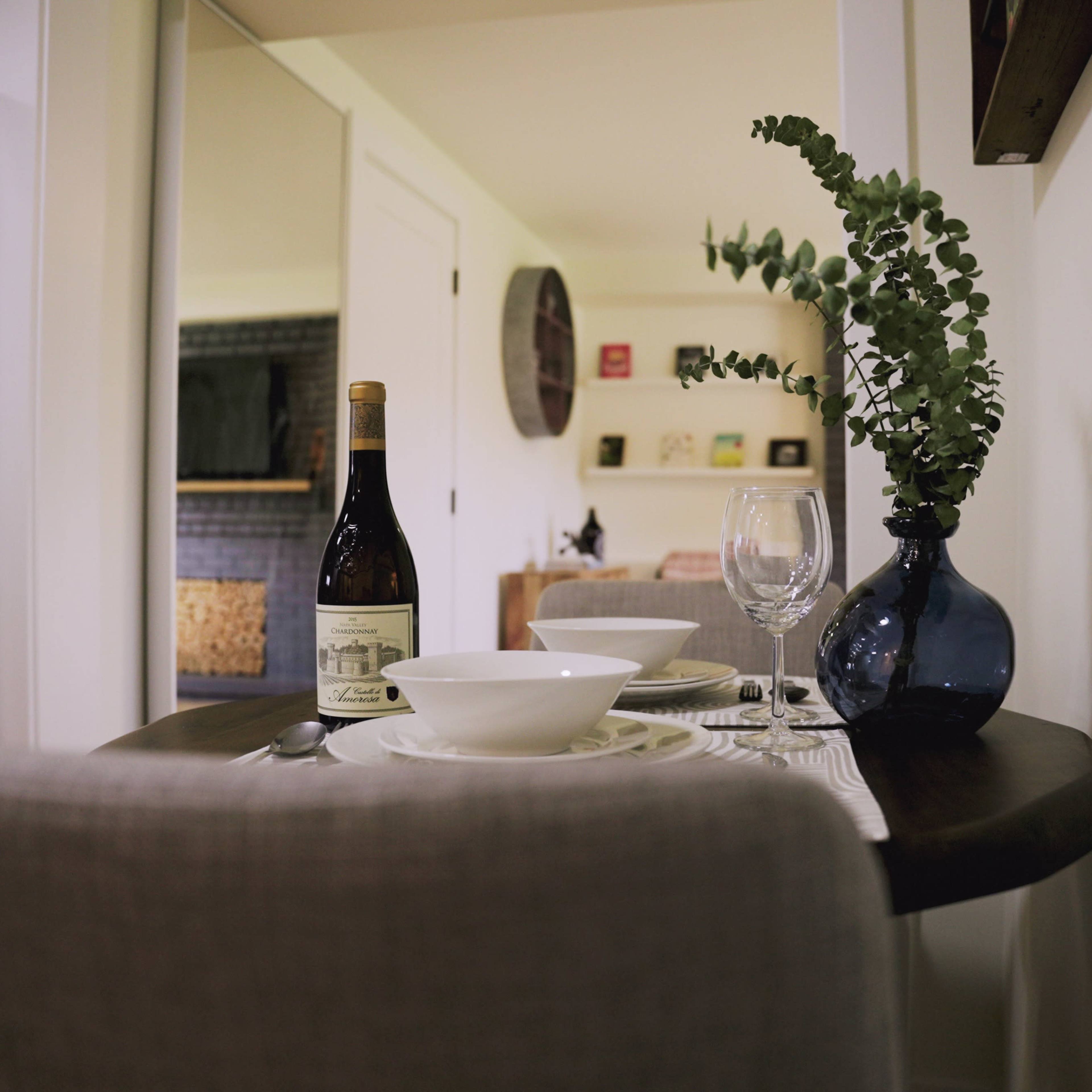 A small dining table is set with a bowl, glassware, and a bottle of wine, accompanied by a vase containing green foliage.
