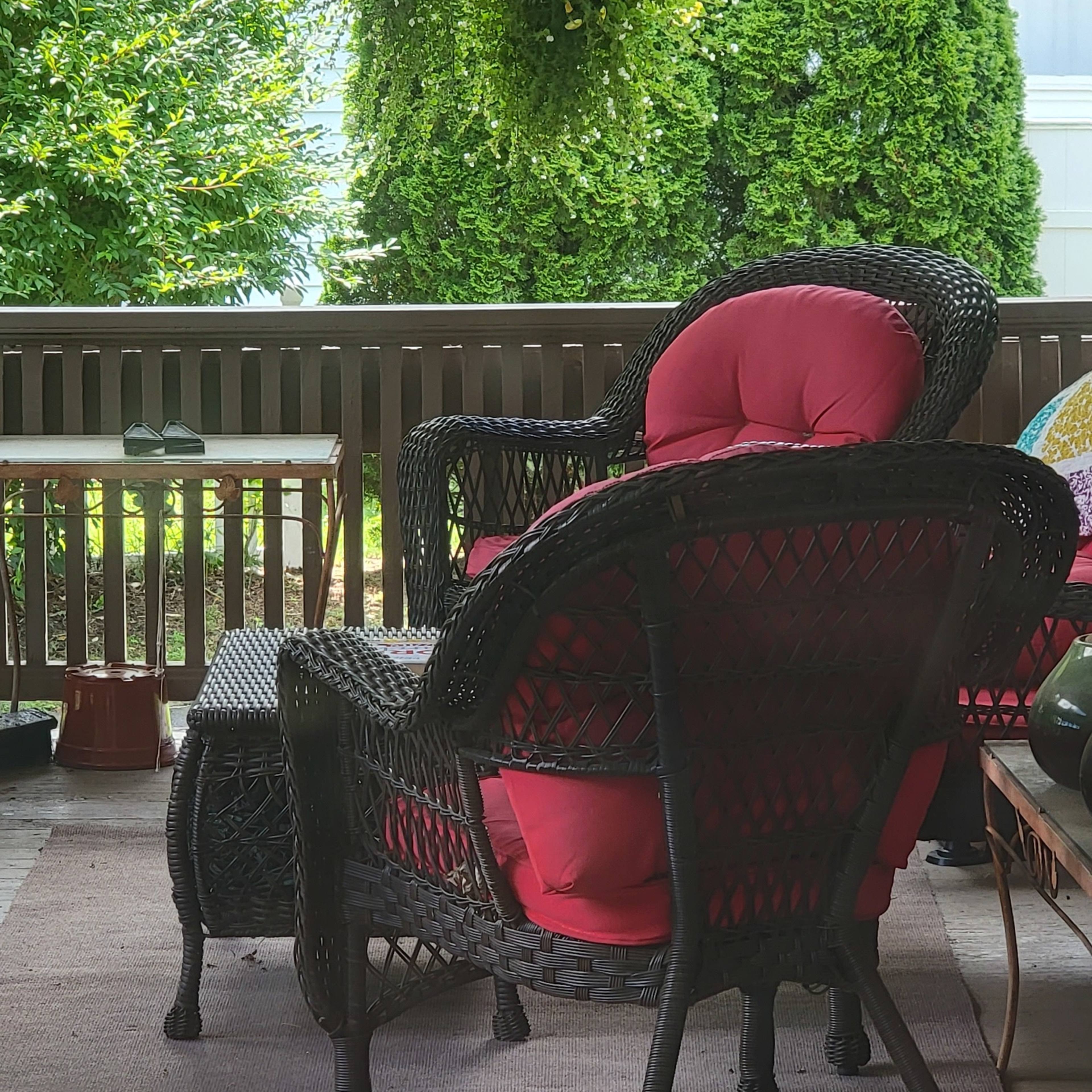 A pair of black wicker chairs with red cushions are positioned on a porch in front of green foliage and a hanging planter.