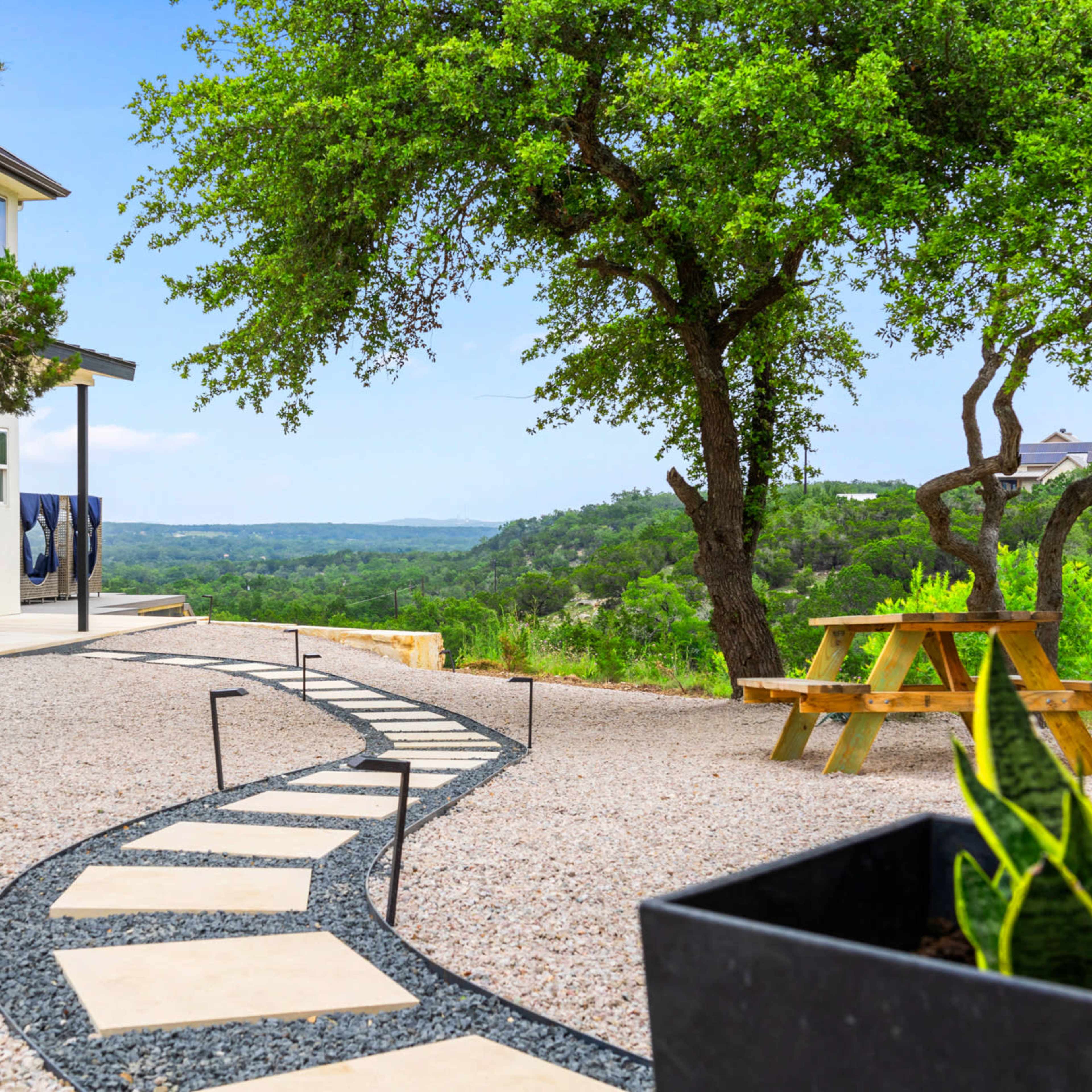 A stone pathway leads from a modern white house through a gravel yard to a wooden picnic table under a tree, with a scenic view of distant hills.