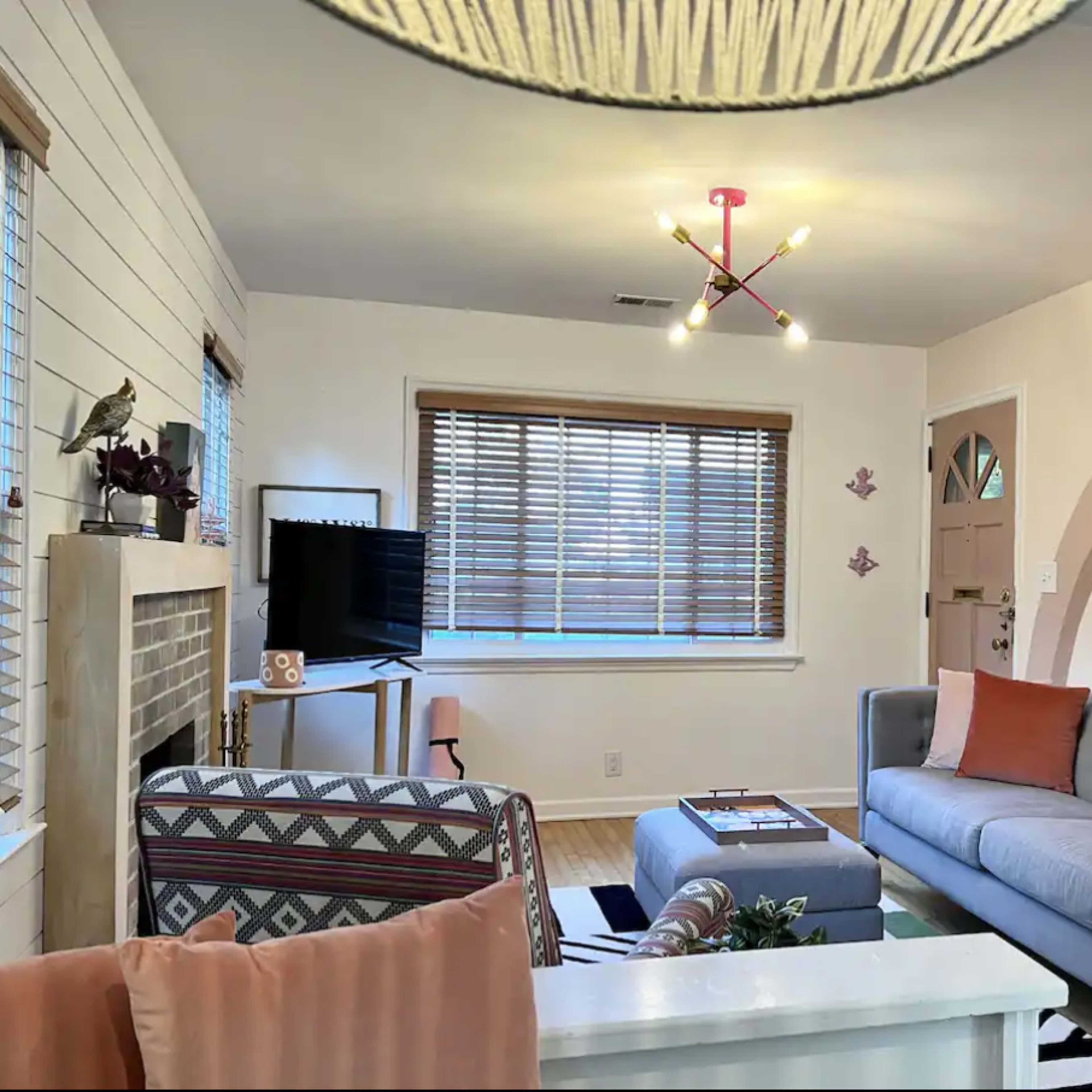 A living room with a gray sofa, a patterned armchair, a television on a stand, and a coffee table, all set against a backdrop of light-colored walls and wooden blinds.
