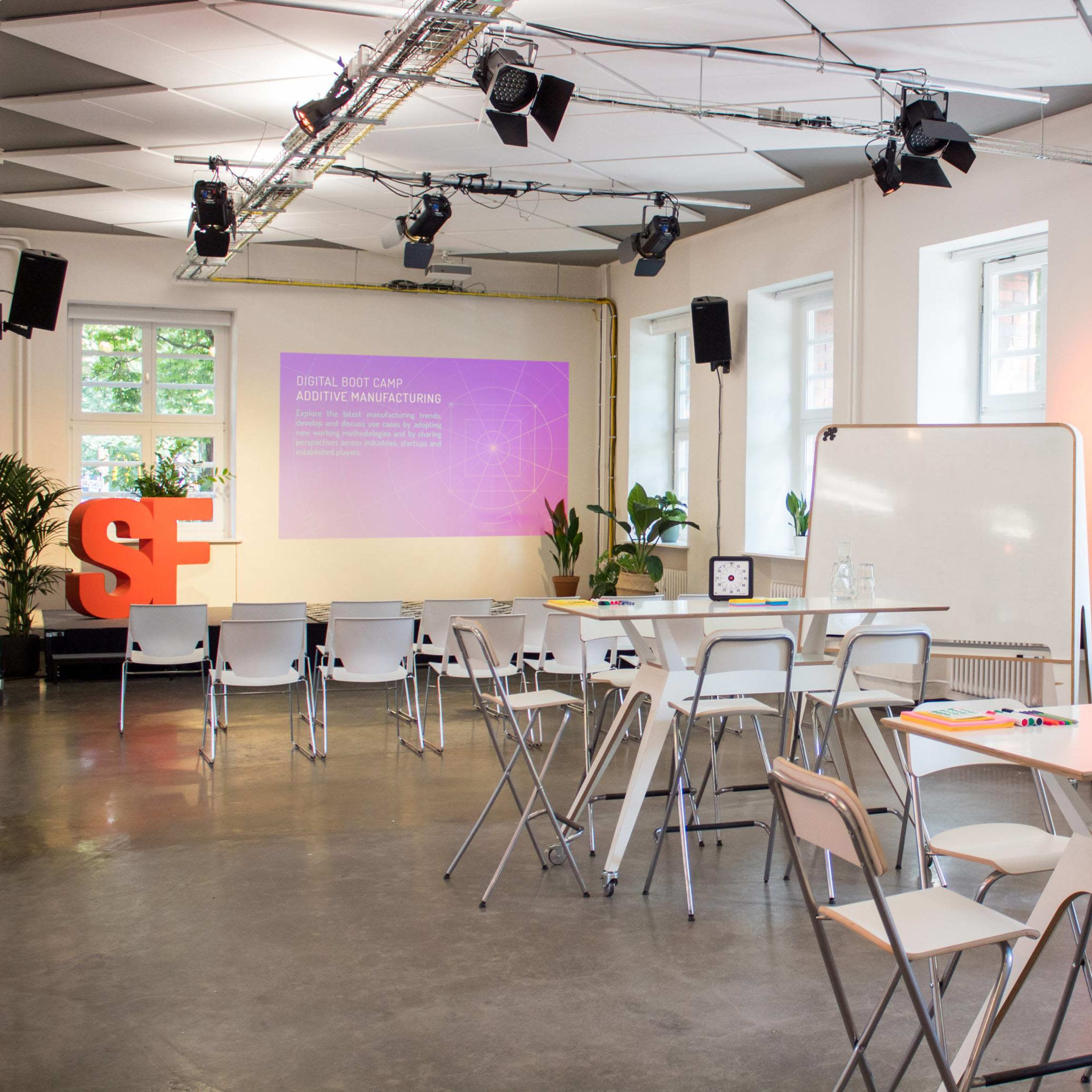 A modern, well-lit seminar room set up for a presentation, featuring chairs, tables, and a large screen displaying a digital presentation.