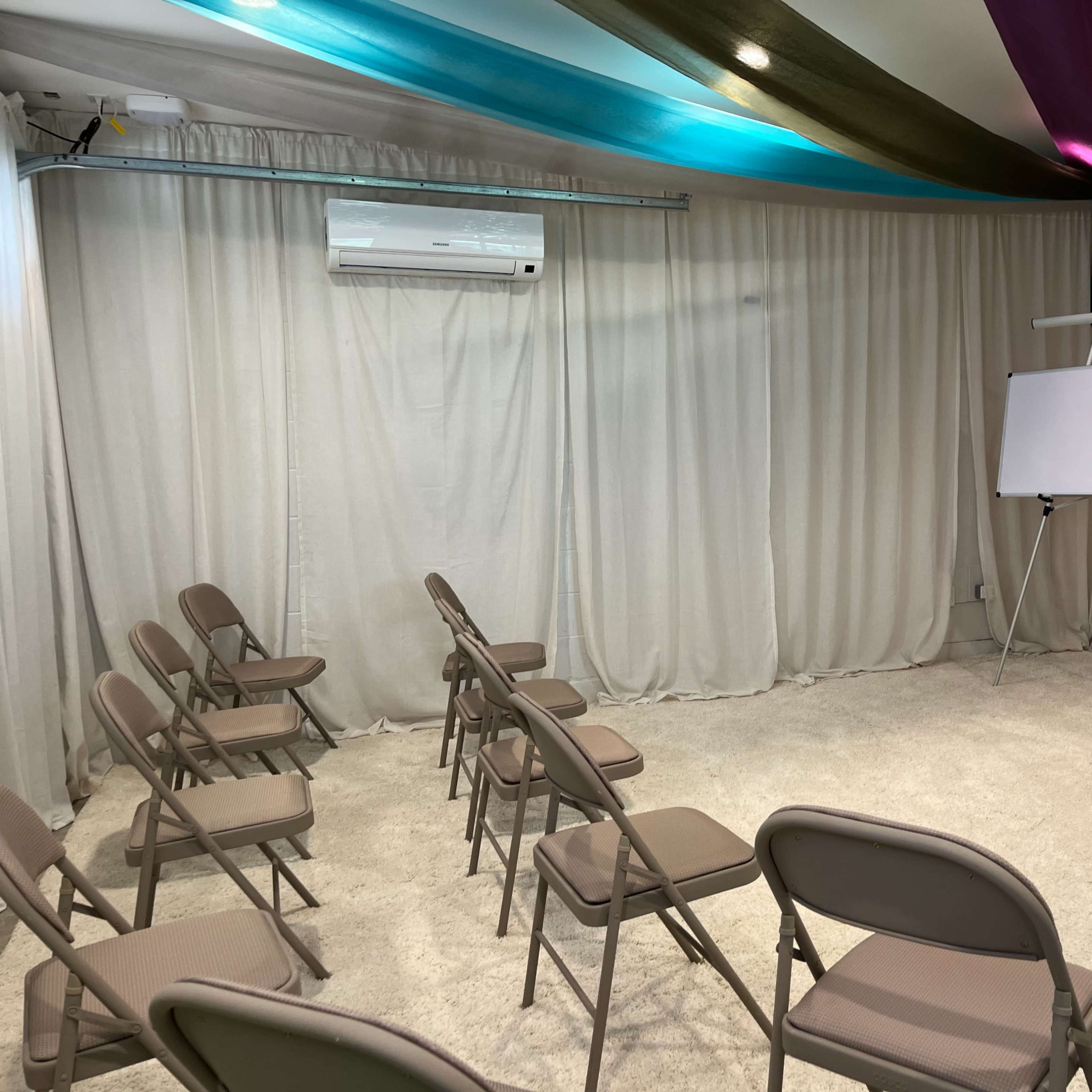 A room with several folding chairs arranged in rows facing a blank whiteboard, with draped curtains and colored fabric overhead.