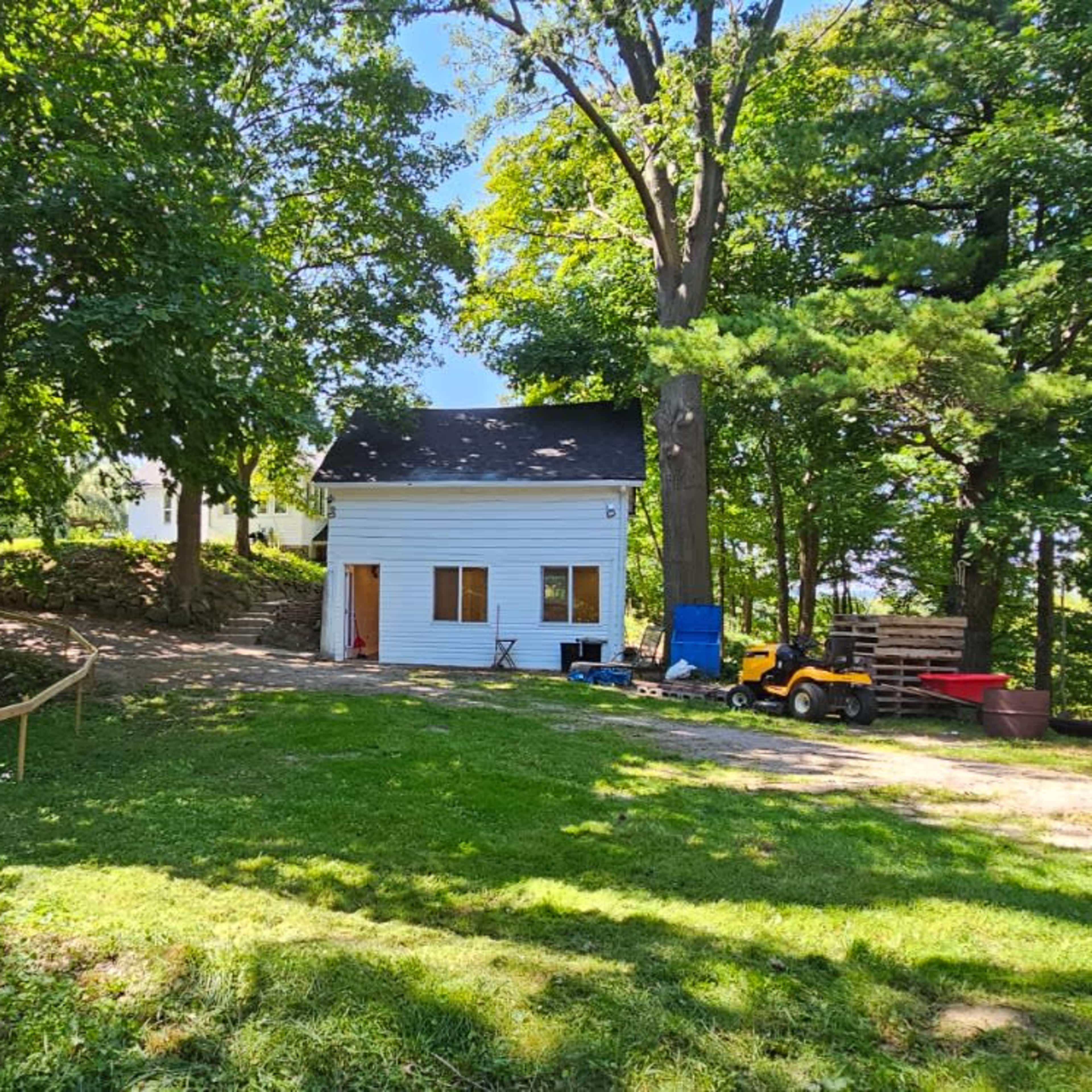 A small white house is situated among trees on a grassy area, with a gravel path leading up to it.