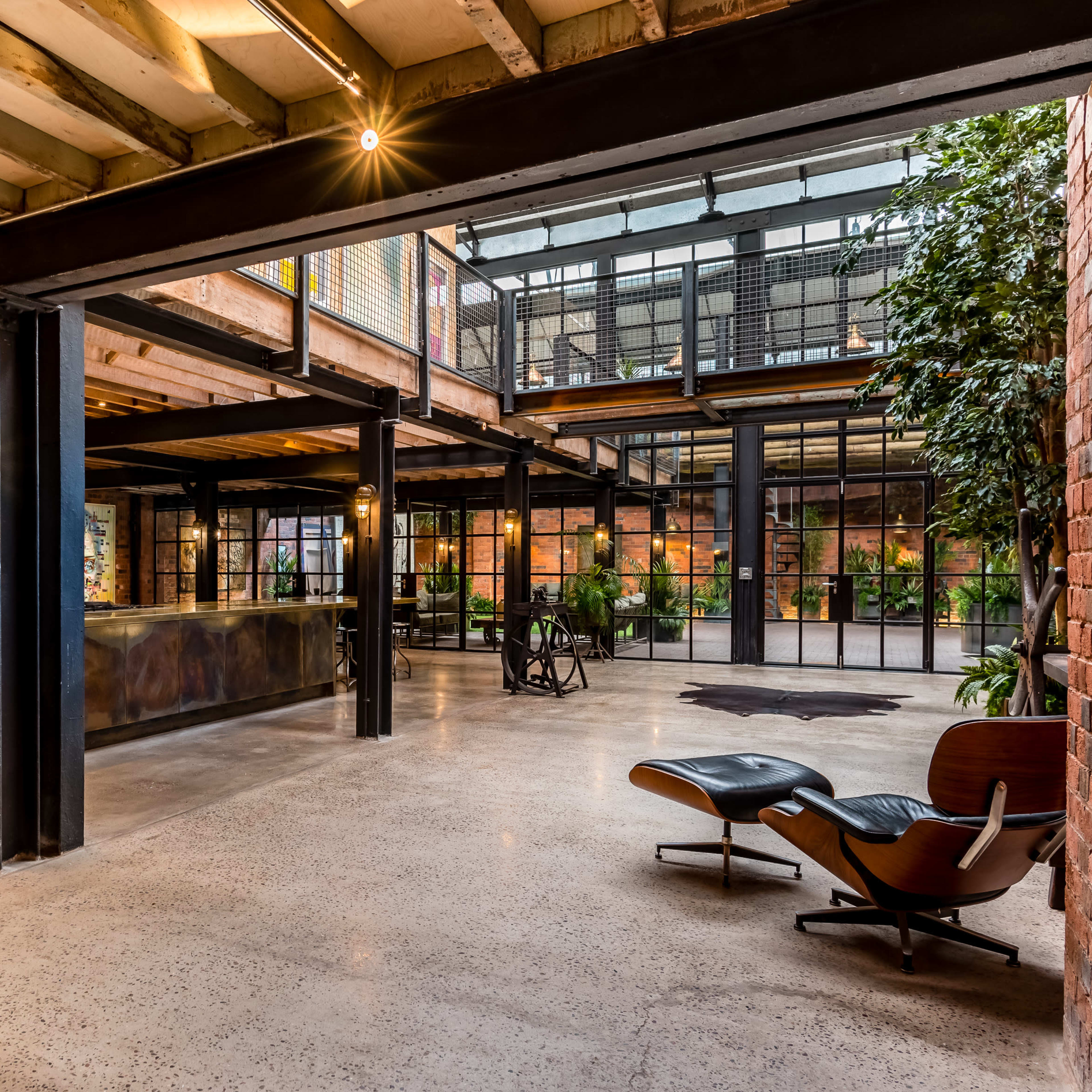 A spacious industrial-style interior with exposed brick walls, large windows, and a mix of modern furniture and plants.