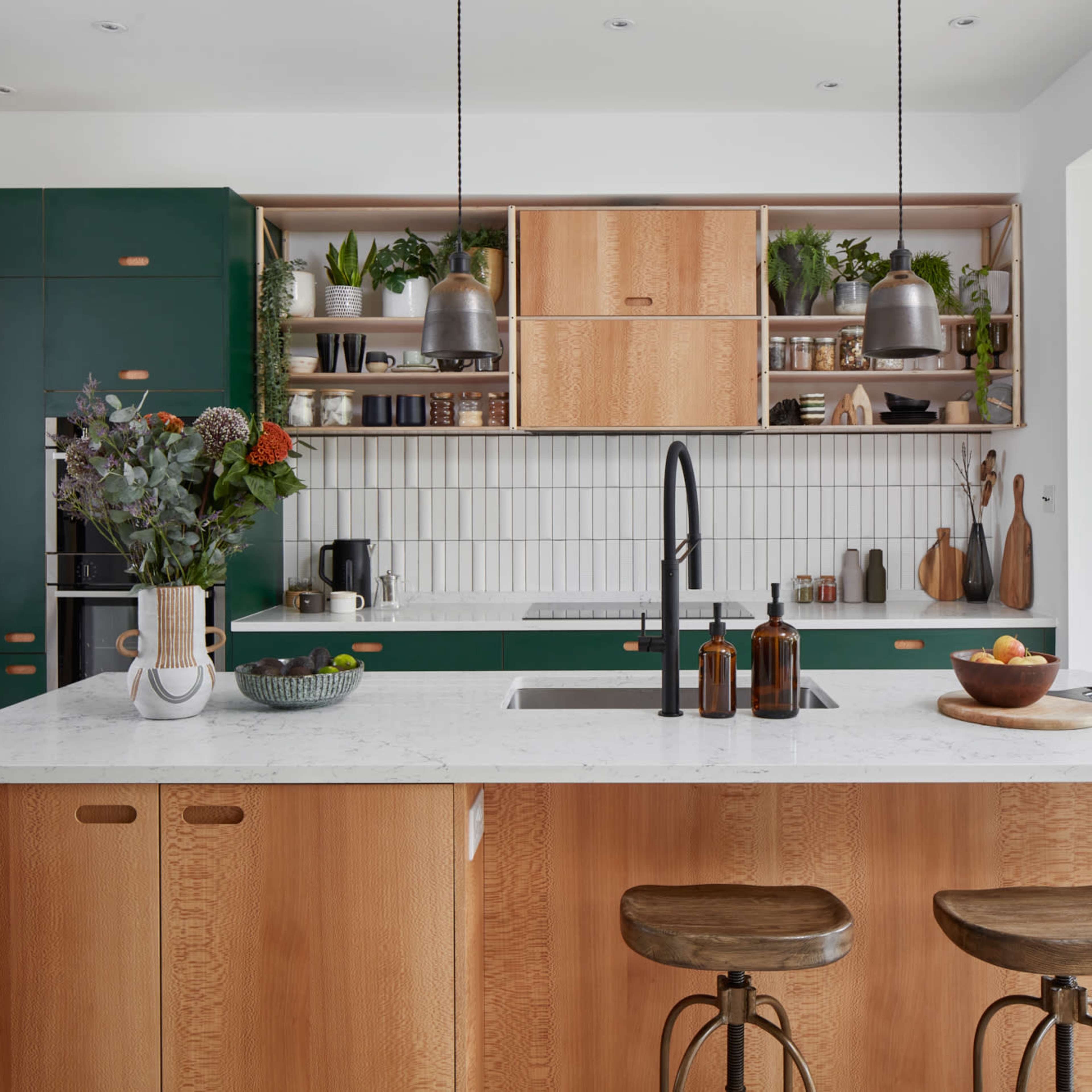 A modern kitchen features green cabinetry, wooden elements, a white countertop, and shelves adorned with plants and kitchenware.