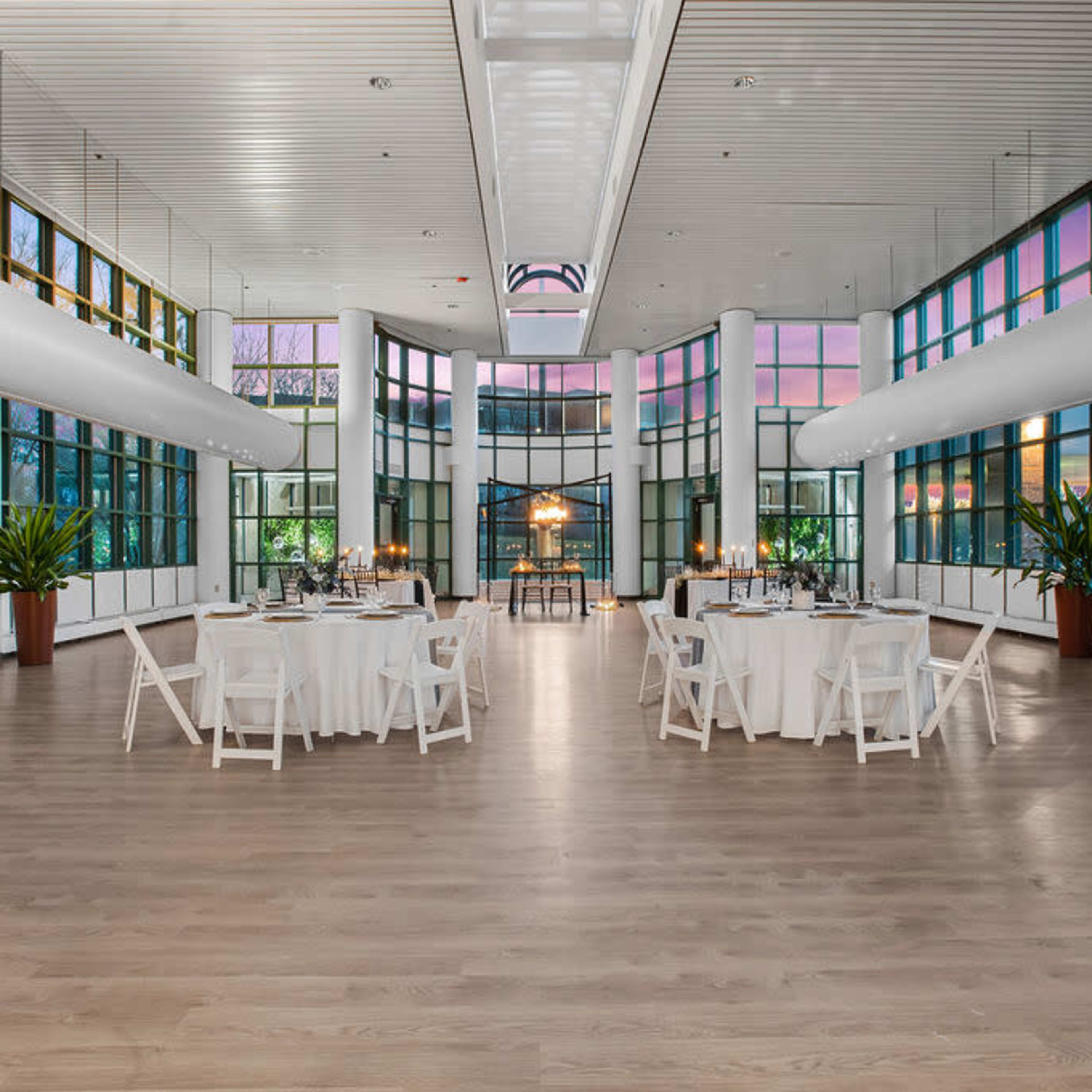 A spacious event room with large windows, featuring white tables set for a gathering and potted plants in the corners.