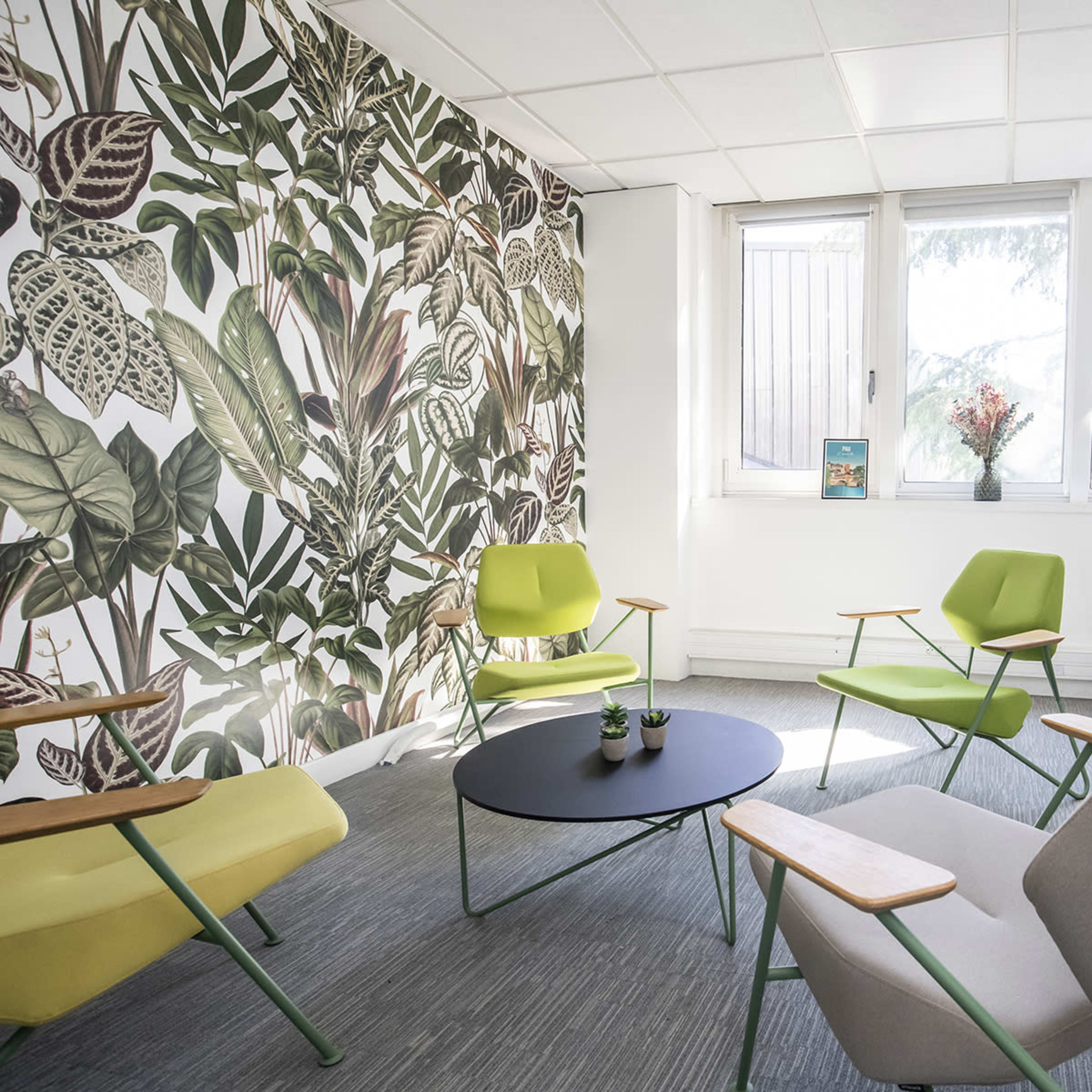 A modern seating area features green and gray chairs arranged around a black coffee table, with a plant-themed wallpaper and large windows letting in natural light.