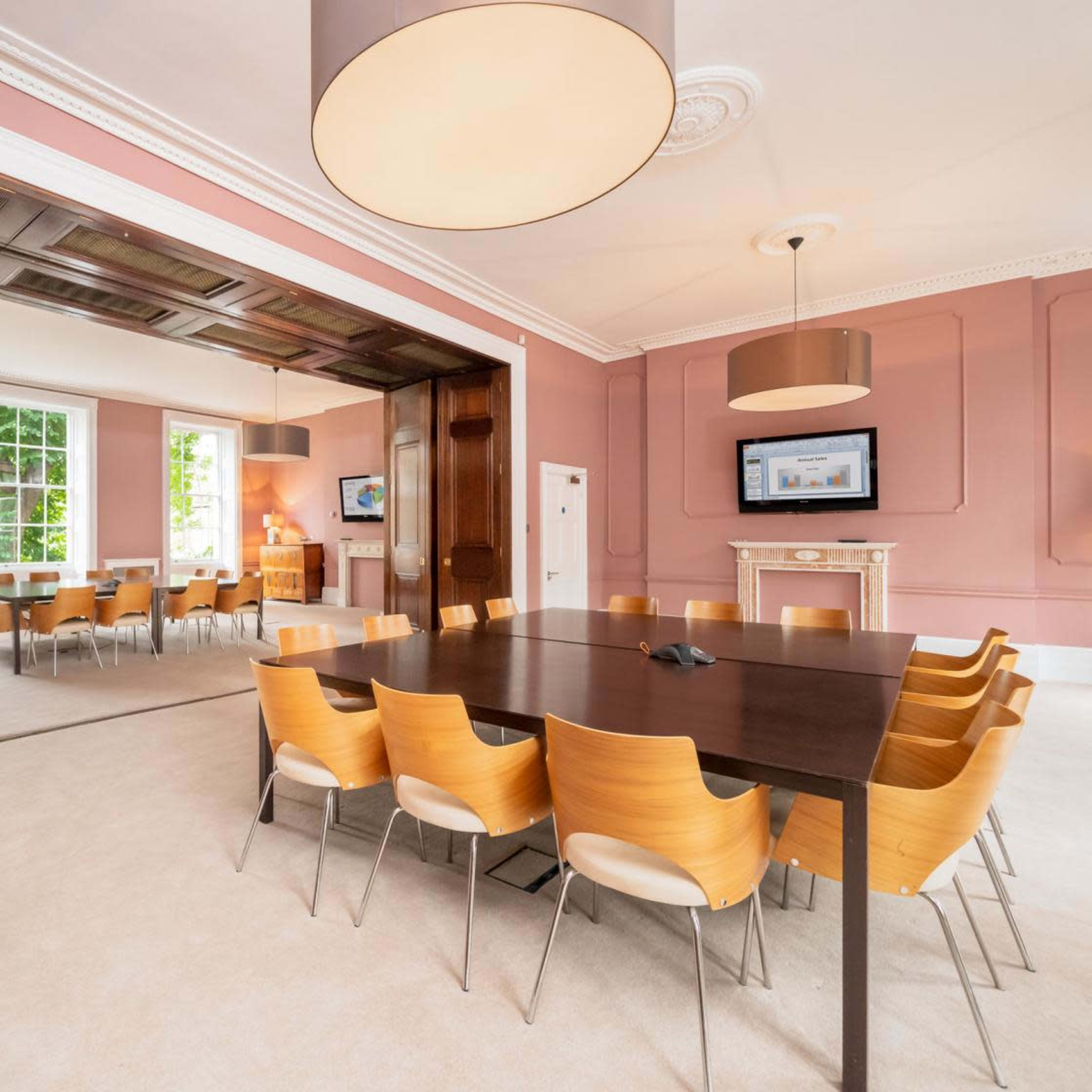 A spacious meeting room features a large wooden table surrounded by eight wooden chairs, with soft natural light coming through large windows.