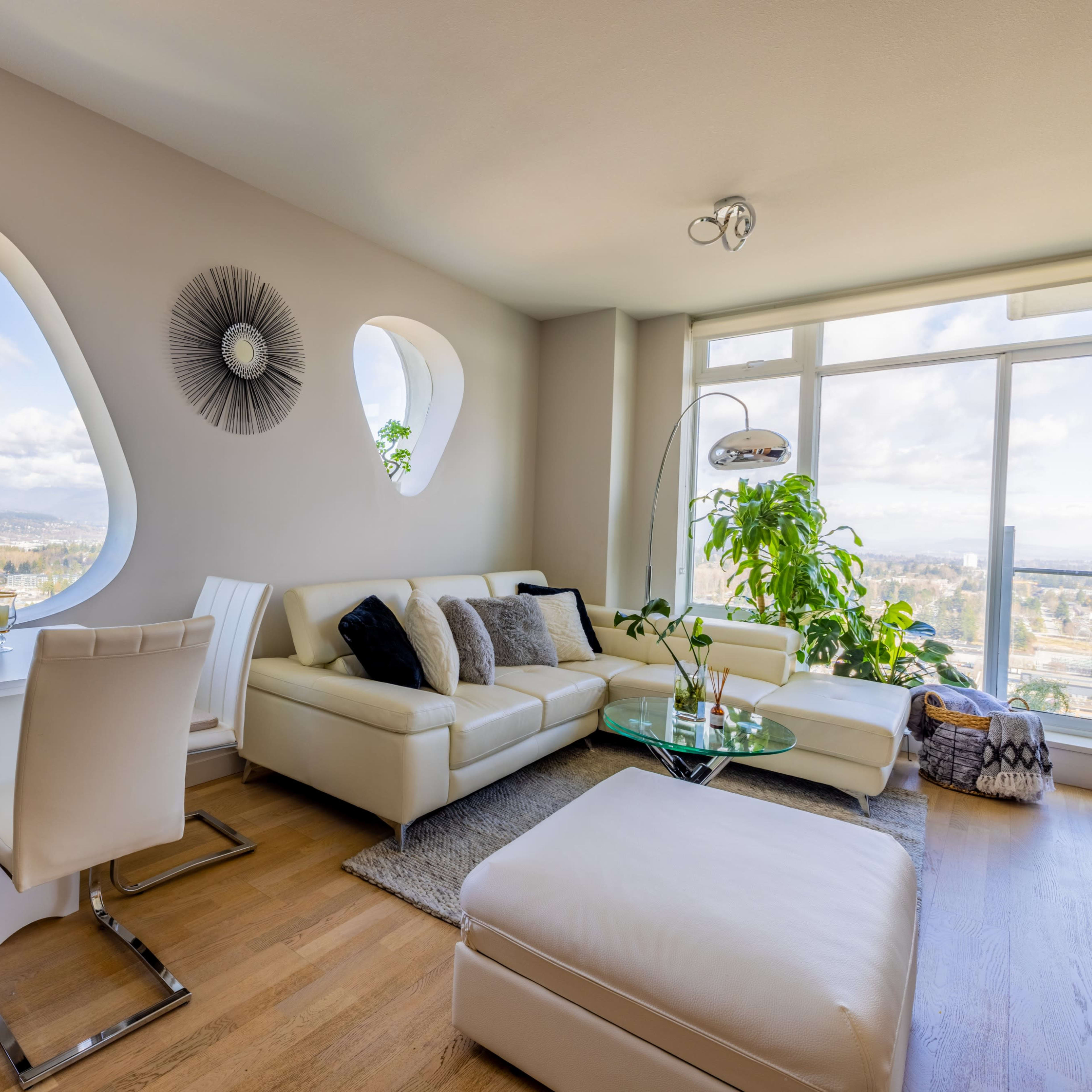A modern living room features a white leather sofa, a glass coffee table, and a dining area with a view through unique, rounded windows.