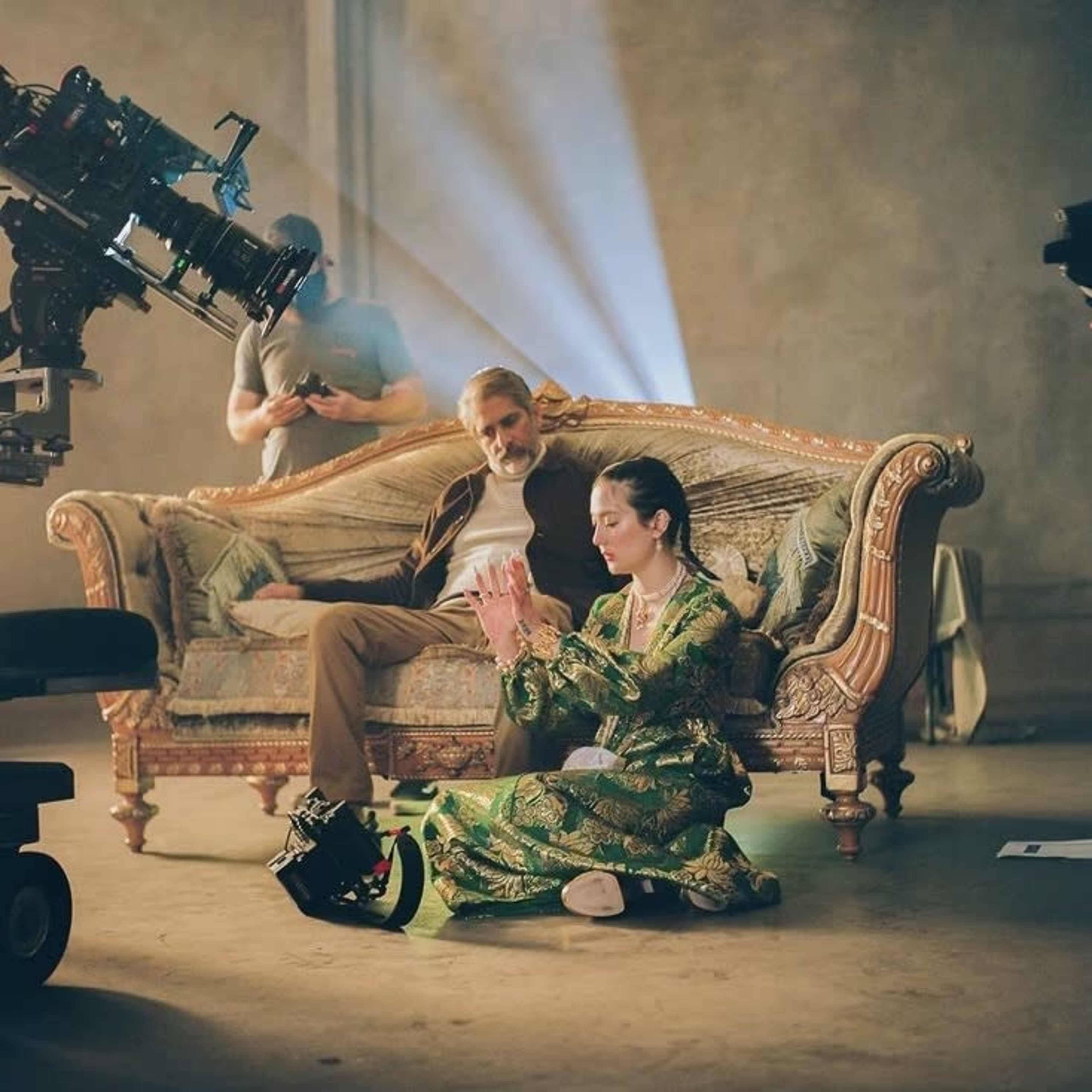 A film crew captures a scene featuring two actors on a vintage sofa, with one actor seated and the other kneeling in a colorful outfit.