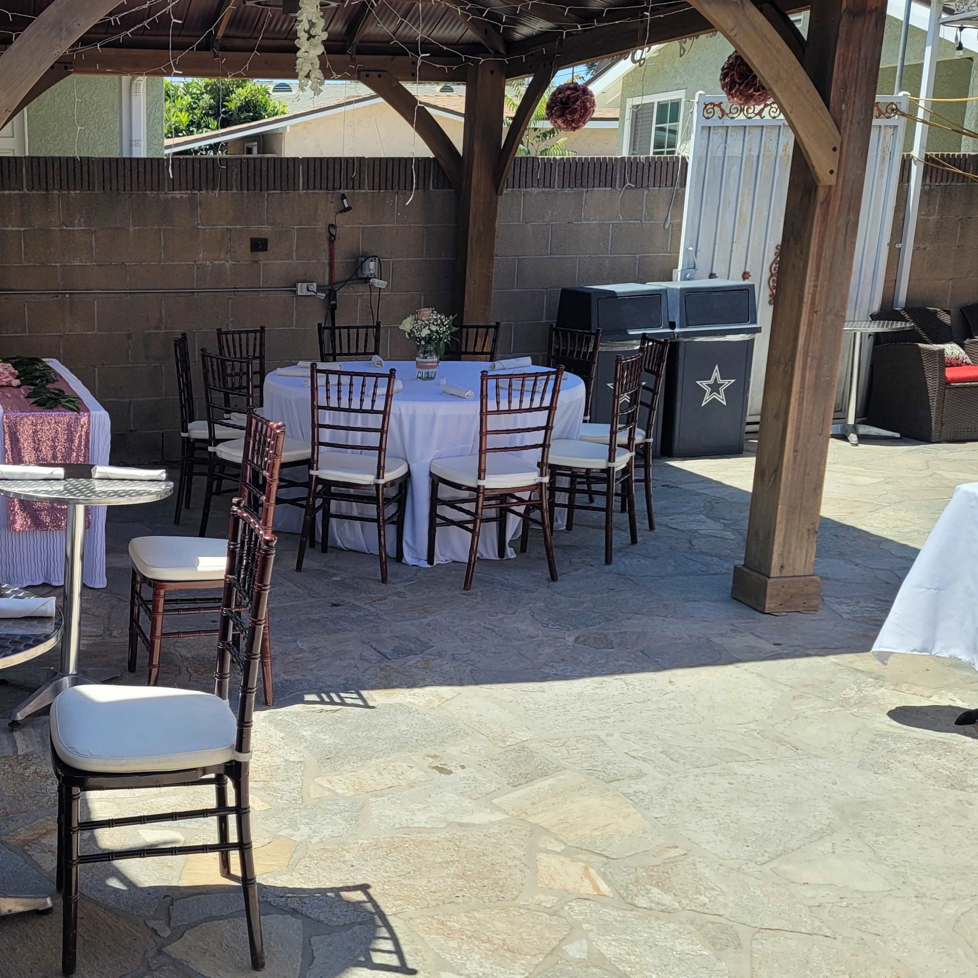 A patio area is set up with several tables and chairs under a wooden gazebo, featuring a stone floor and various decorations.