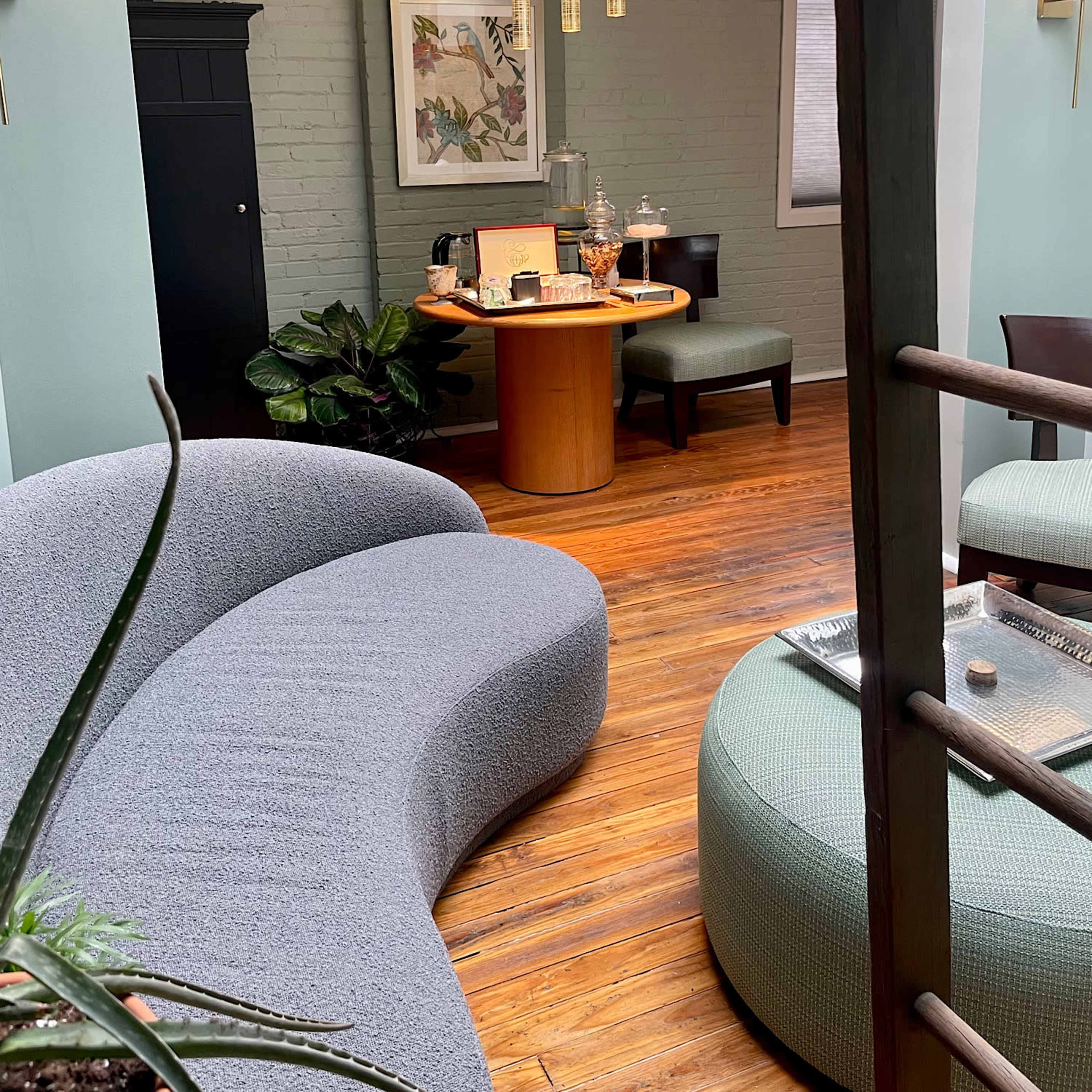 A cozy seating area featuring a curved gray sofa, a round wooden table with refreshments, and green plants against a light-colored wall.