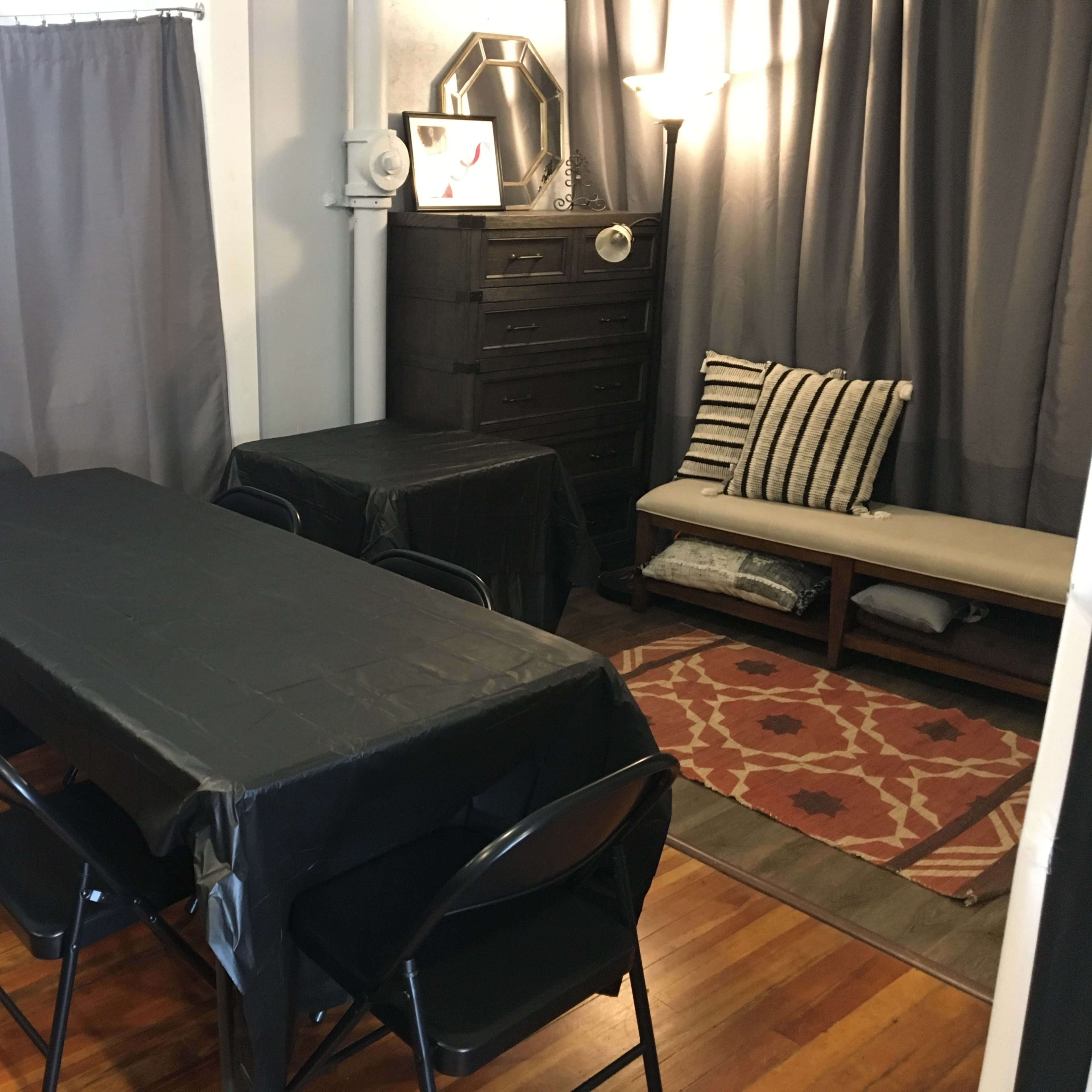 A dining area with a long black table surrounded by folding chairs, a dresser, and a bench with pillows, all set against gray curtains and wooden flooring.