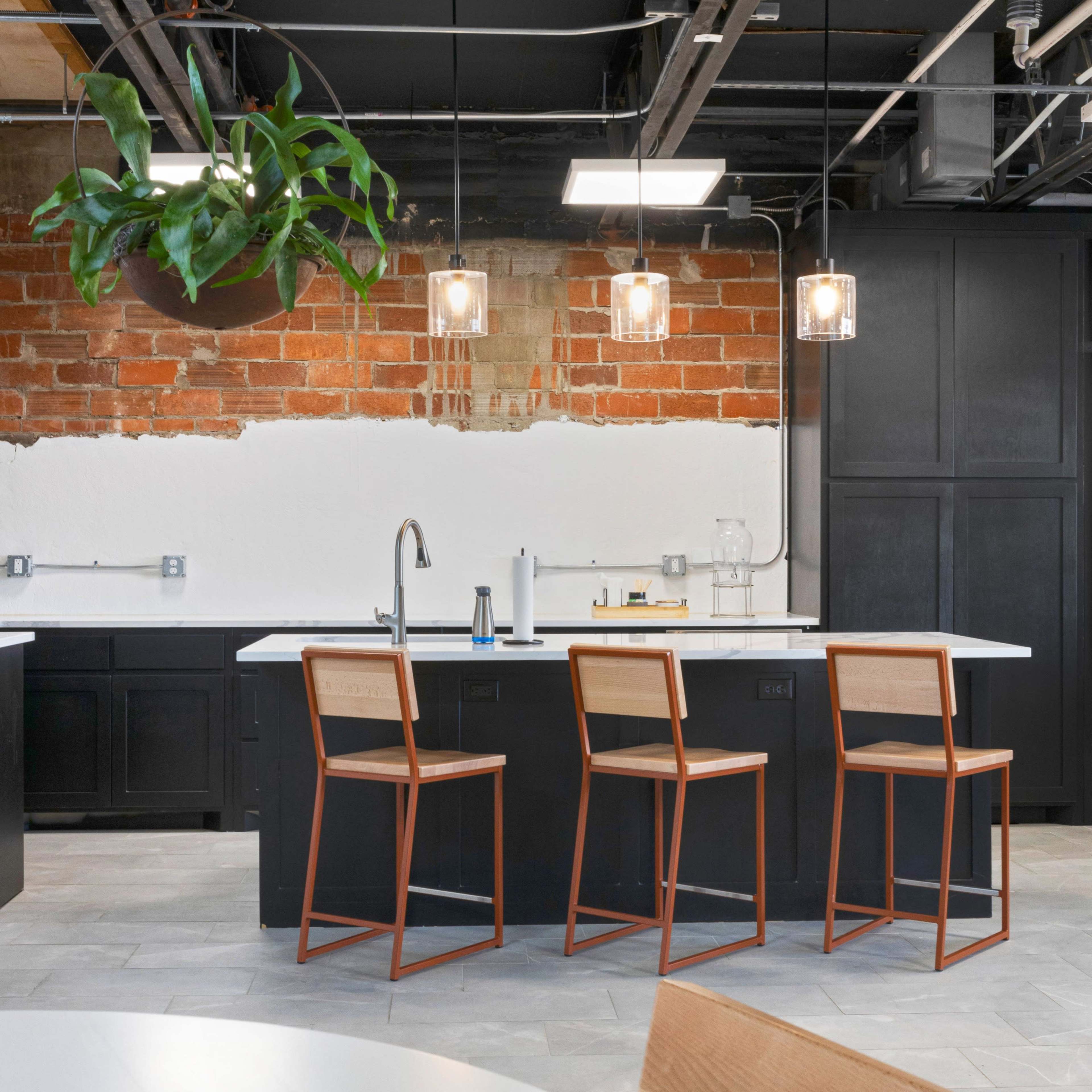 A modern kitchen with a large island, three wooden bar stools, and overhead pendant lights, featuring exposed brick walls and sleek cabinetry.