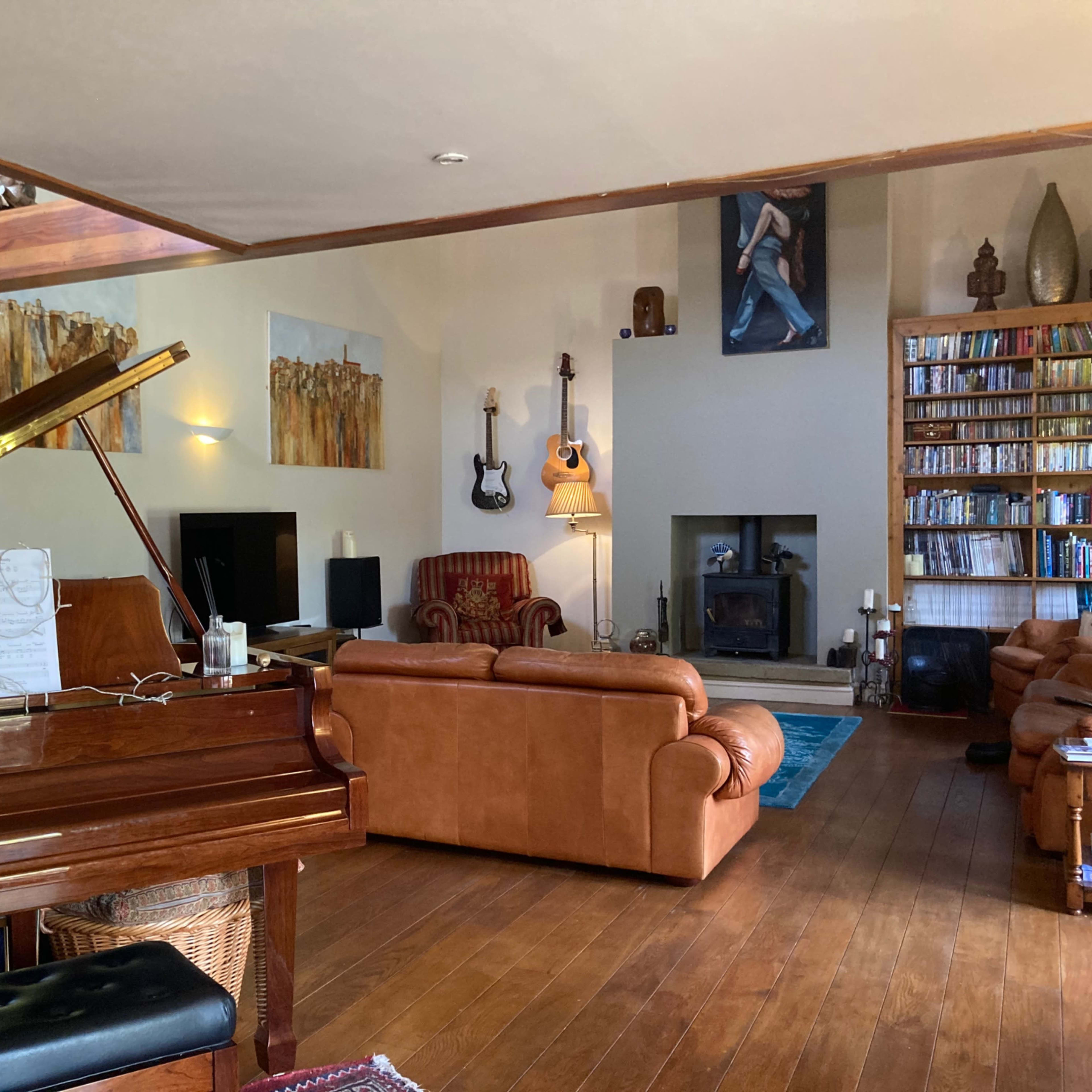 The room features a piano, a bookshelf filled with books, a guitar hanging on the wall, and leather furniture arranged around a central fireplace.