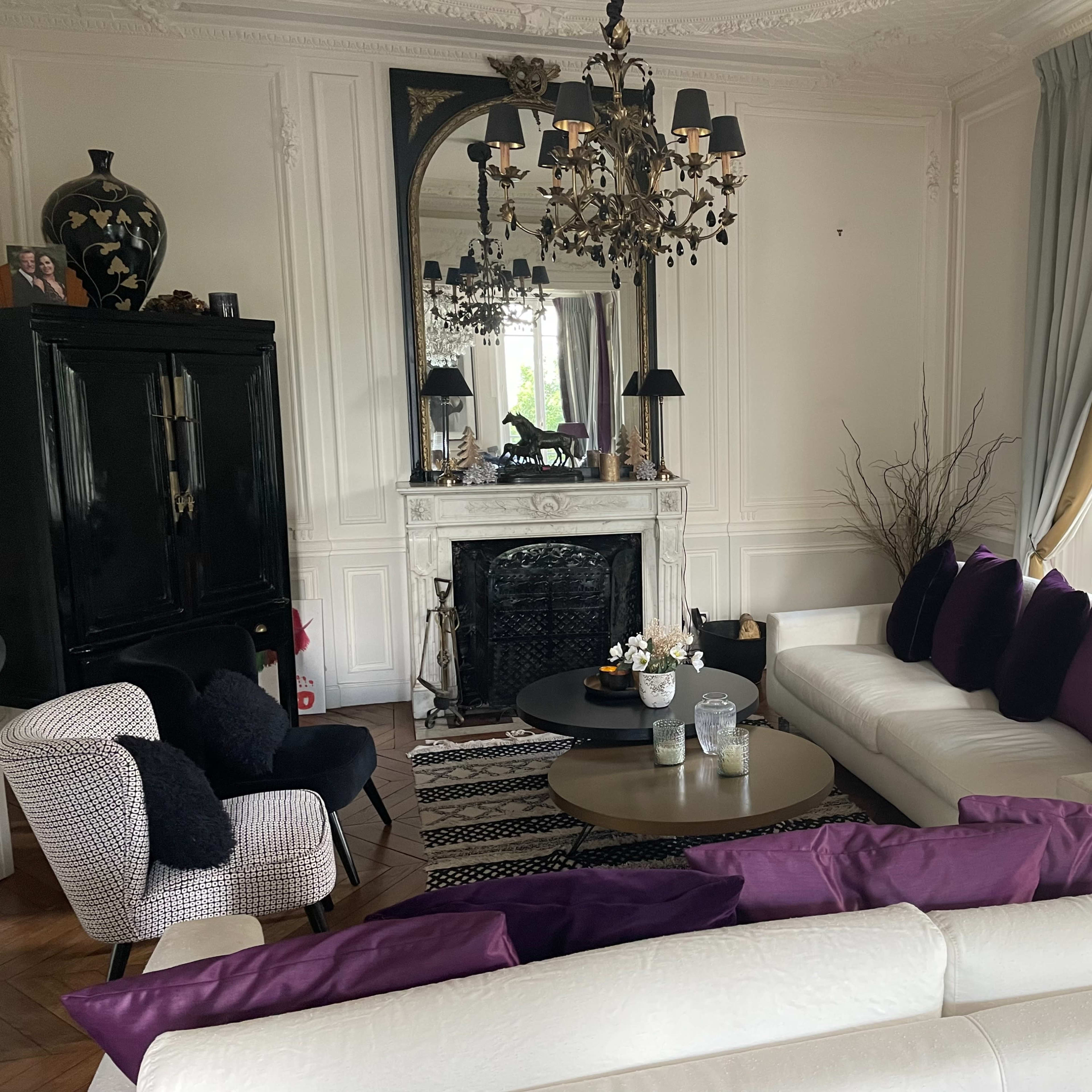 A elegantly styled living room featuring a white sofa with purple cushions, a marble fireplace, a large mirror, and a black side table.