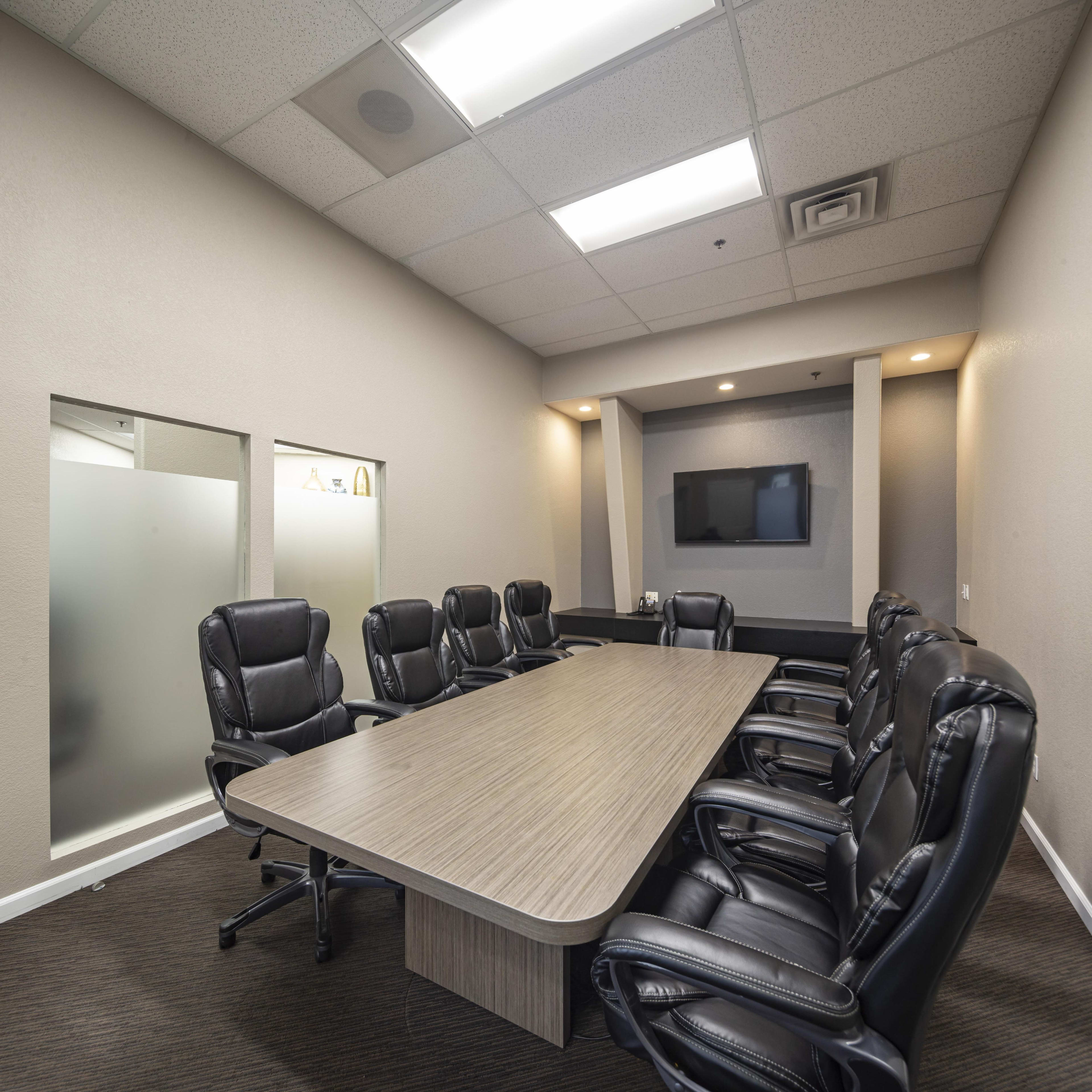 A modern conference room features a long wooden table surrounded by black leather chairs and a wall-mounted TV.