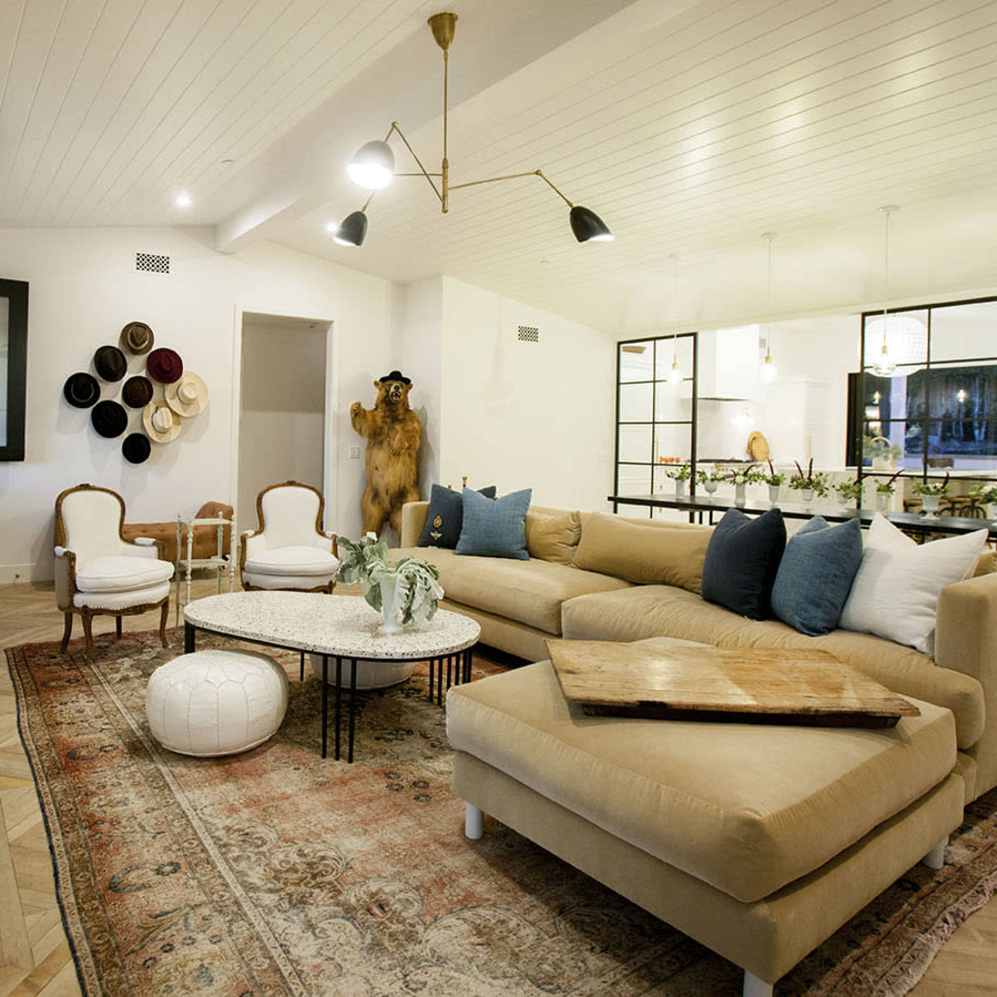 The living room features a large beige sectional couch, a coffee table, and decorative chairs arranged on a patterned rug beneath a paneled ceiling.