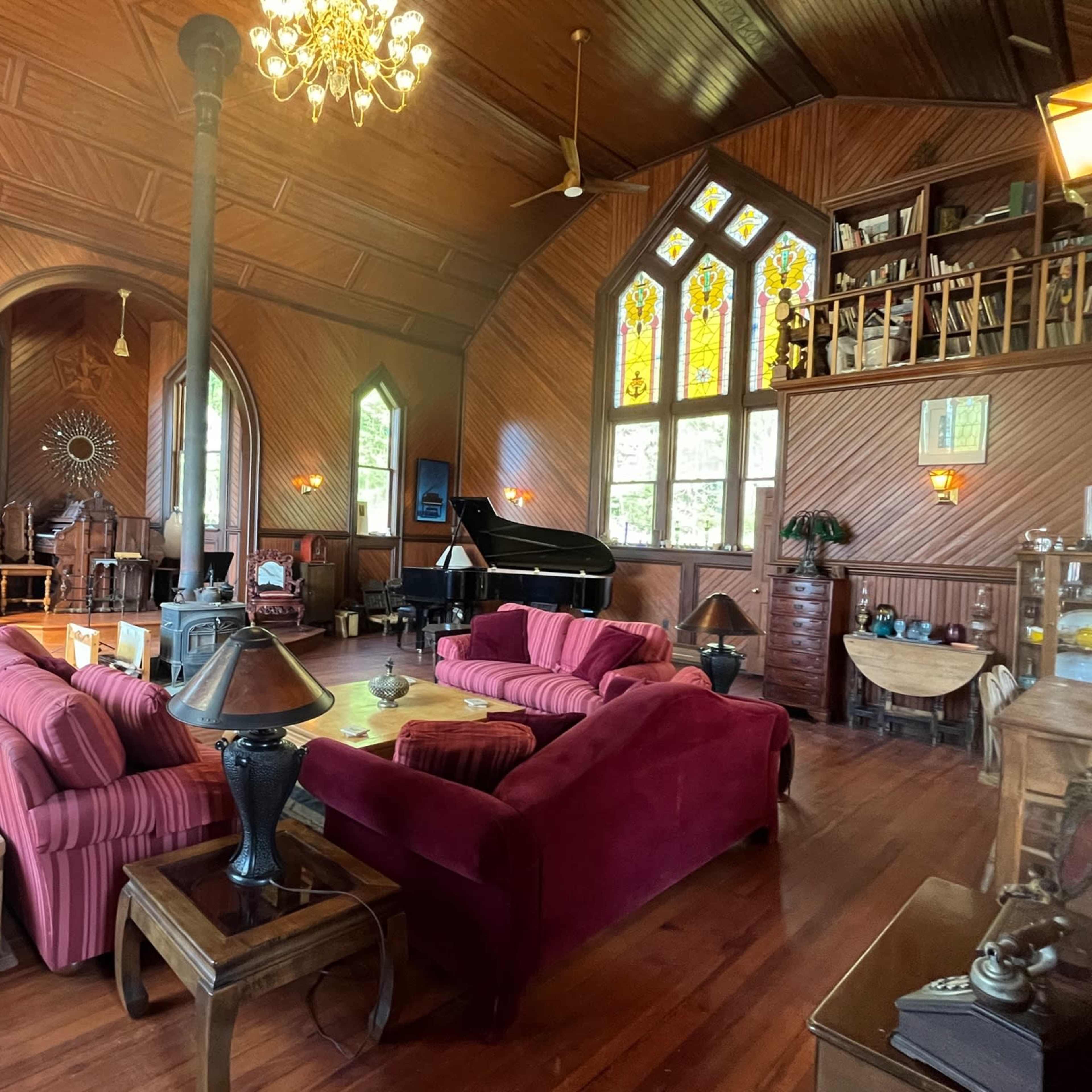 The room features a large, wood-paneled space with a grand piano, stained glass windows, and a mix of vintage furniture arranged around a central seating area.