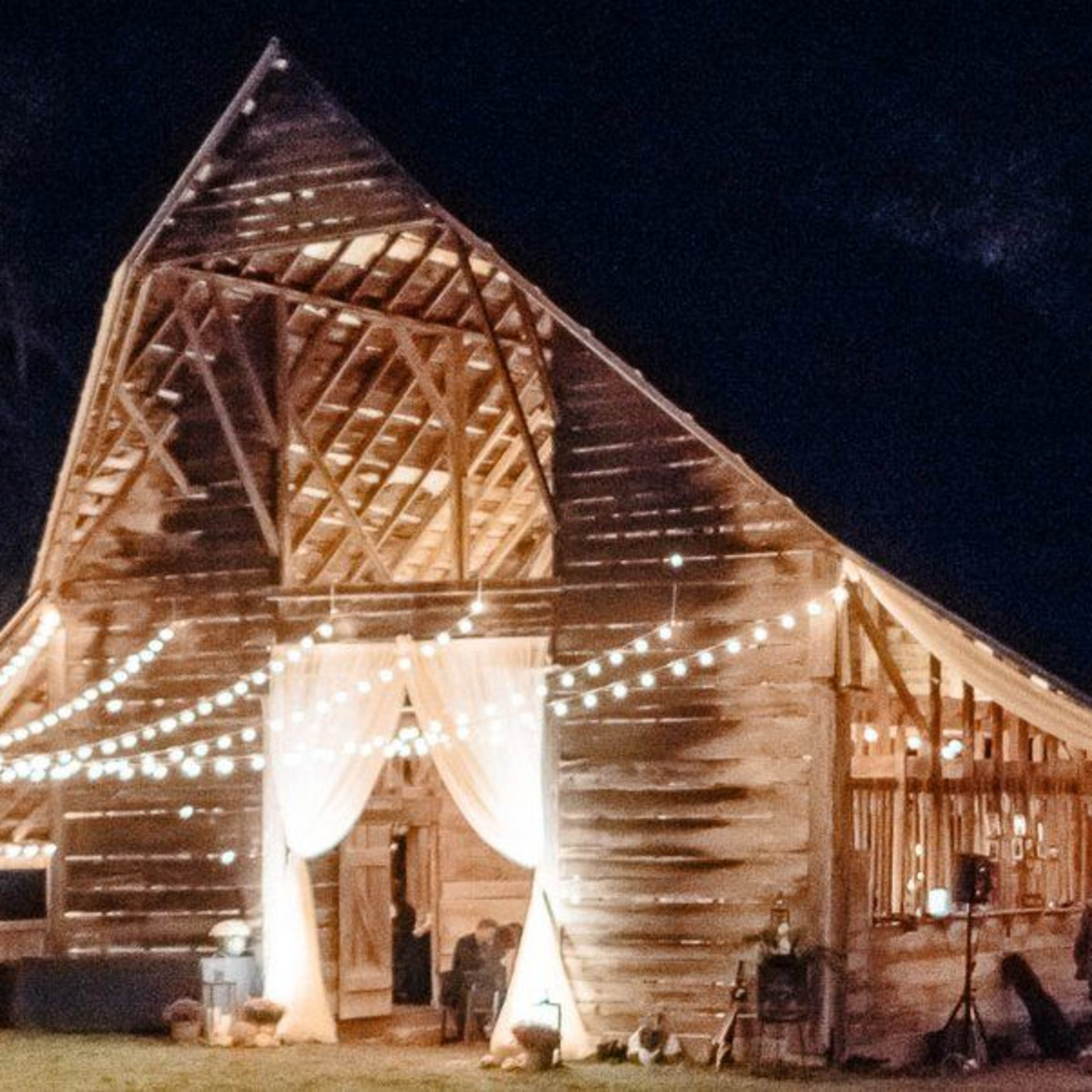 A barn is illuminated at night with string lights and curtains, creating a festive atmosphere.