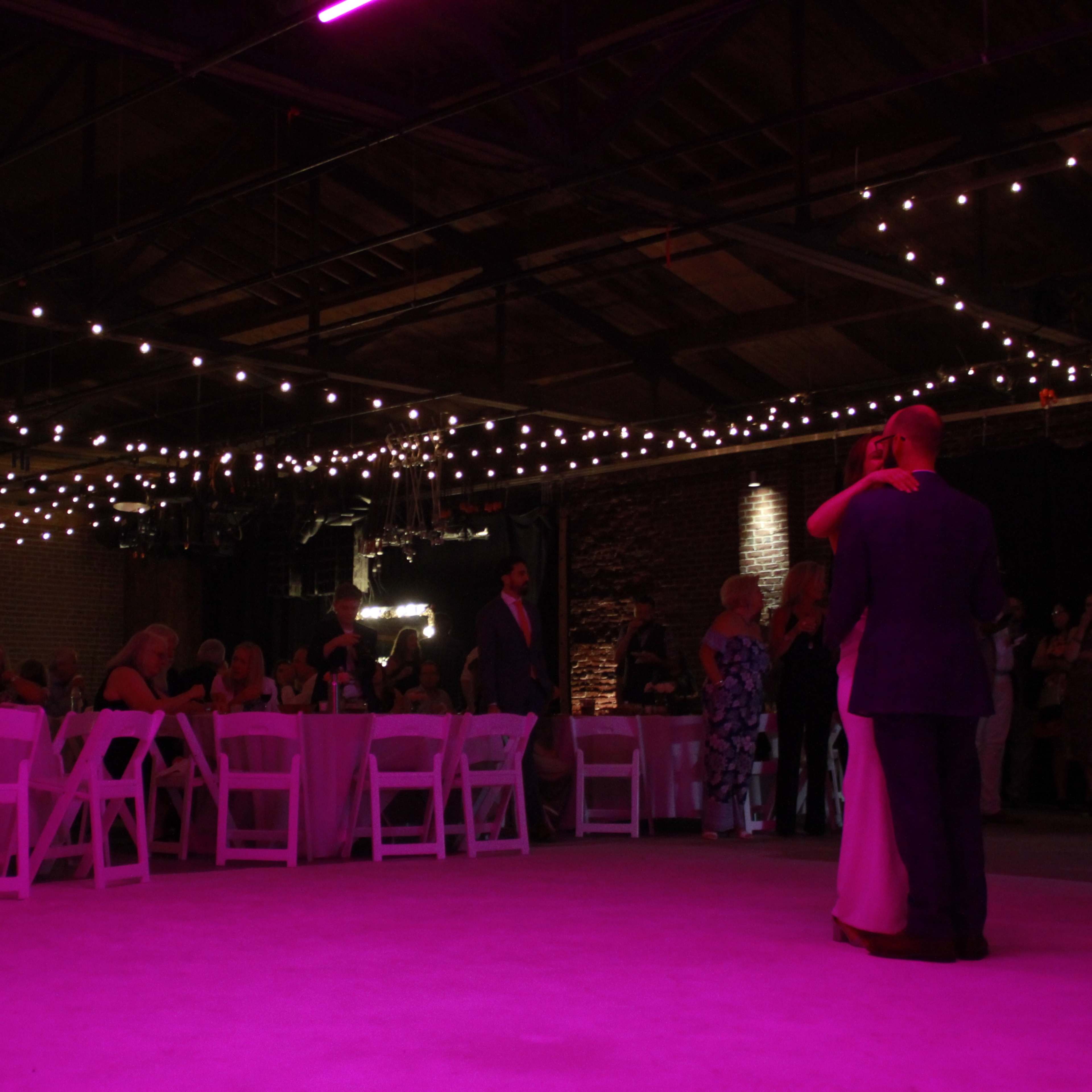 A couple dances on a lighted floor beneath strings of lights in a large indoor event space while guests sit at round tables.