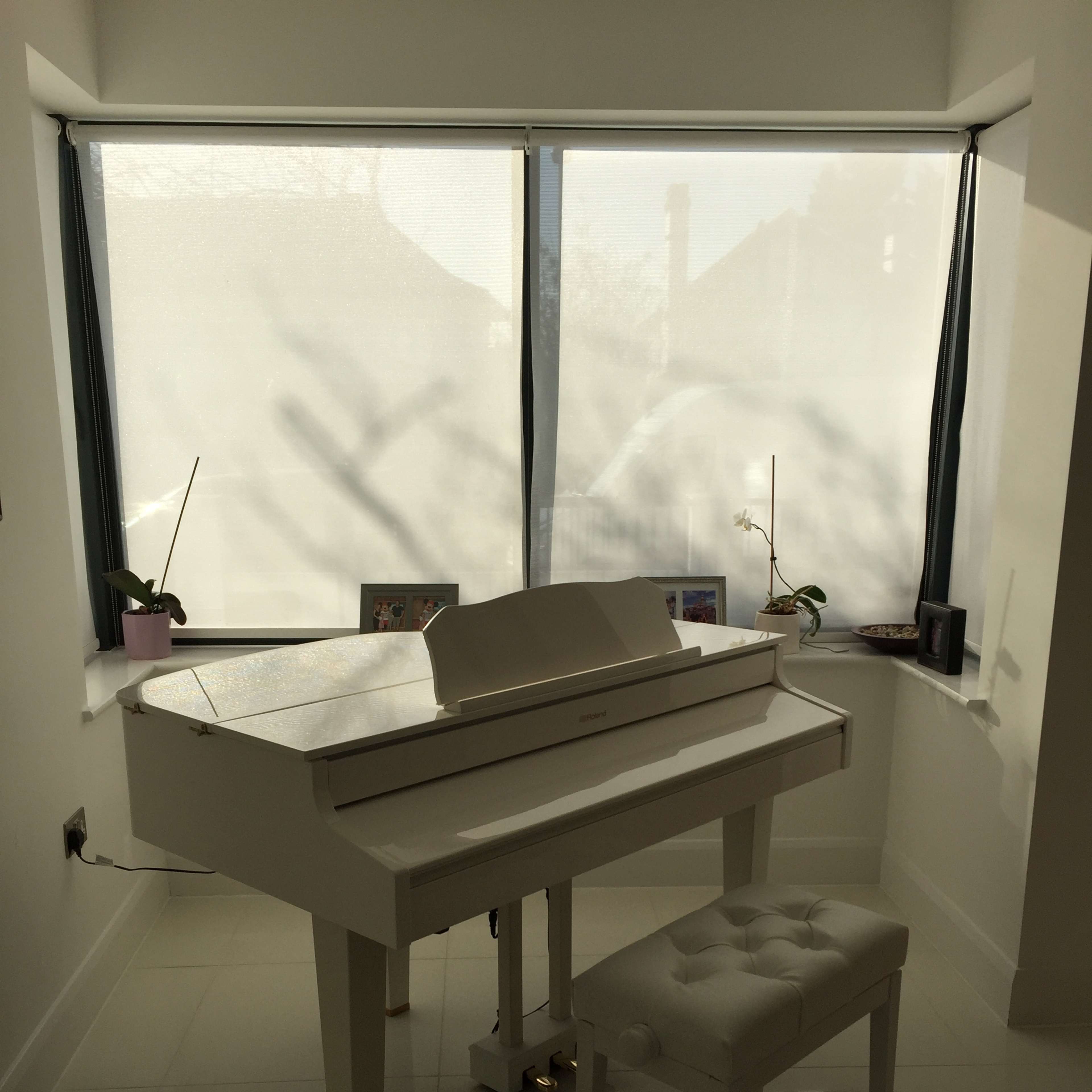 A white piano and matching stool are positioned near a window with sheer curtains, surrounded by photographs and decorative plants.