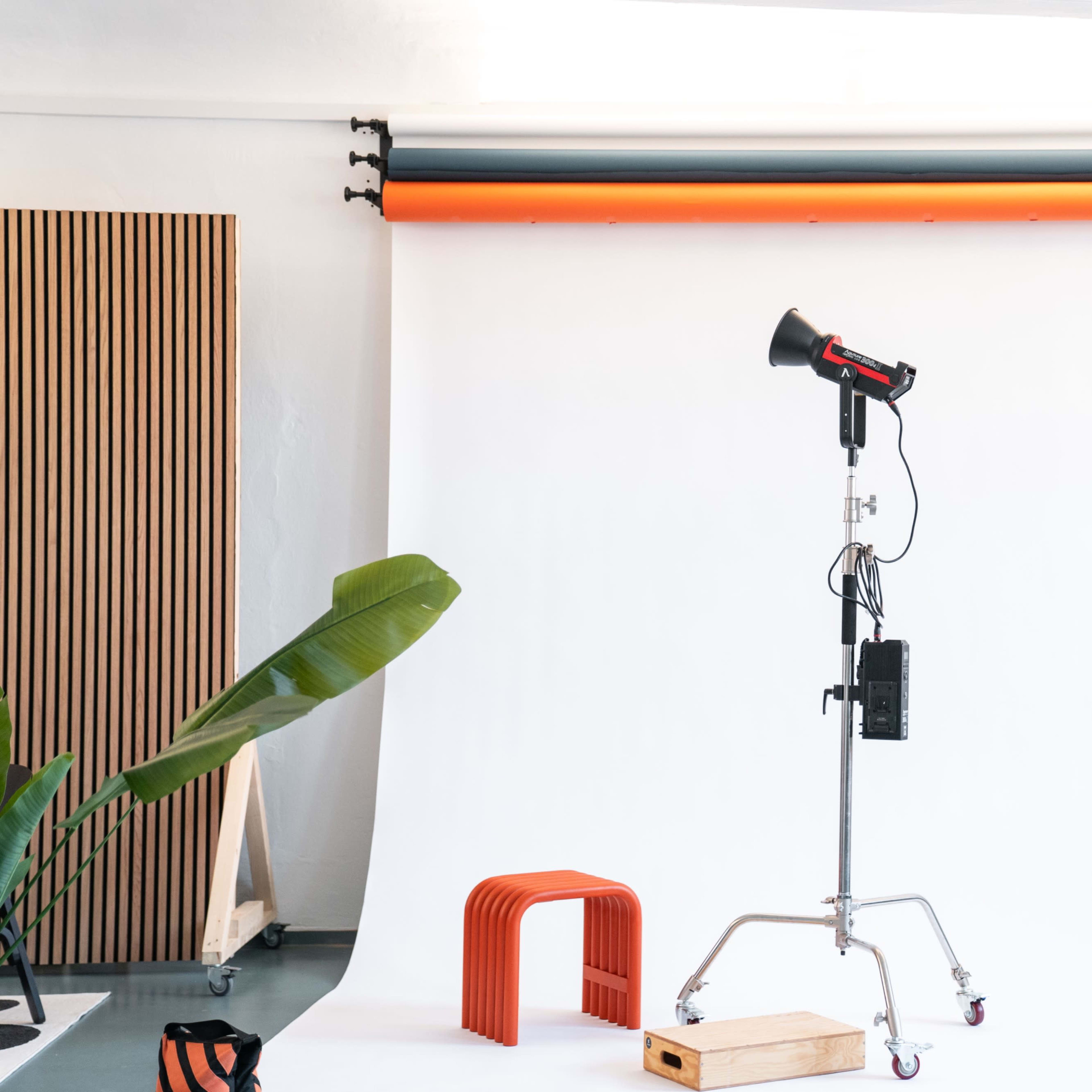 A photography studio setup features a backdrop, a spotlight on a stand, a wooden platform, and a few props, including an orange stool and a hanging rack.