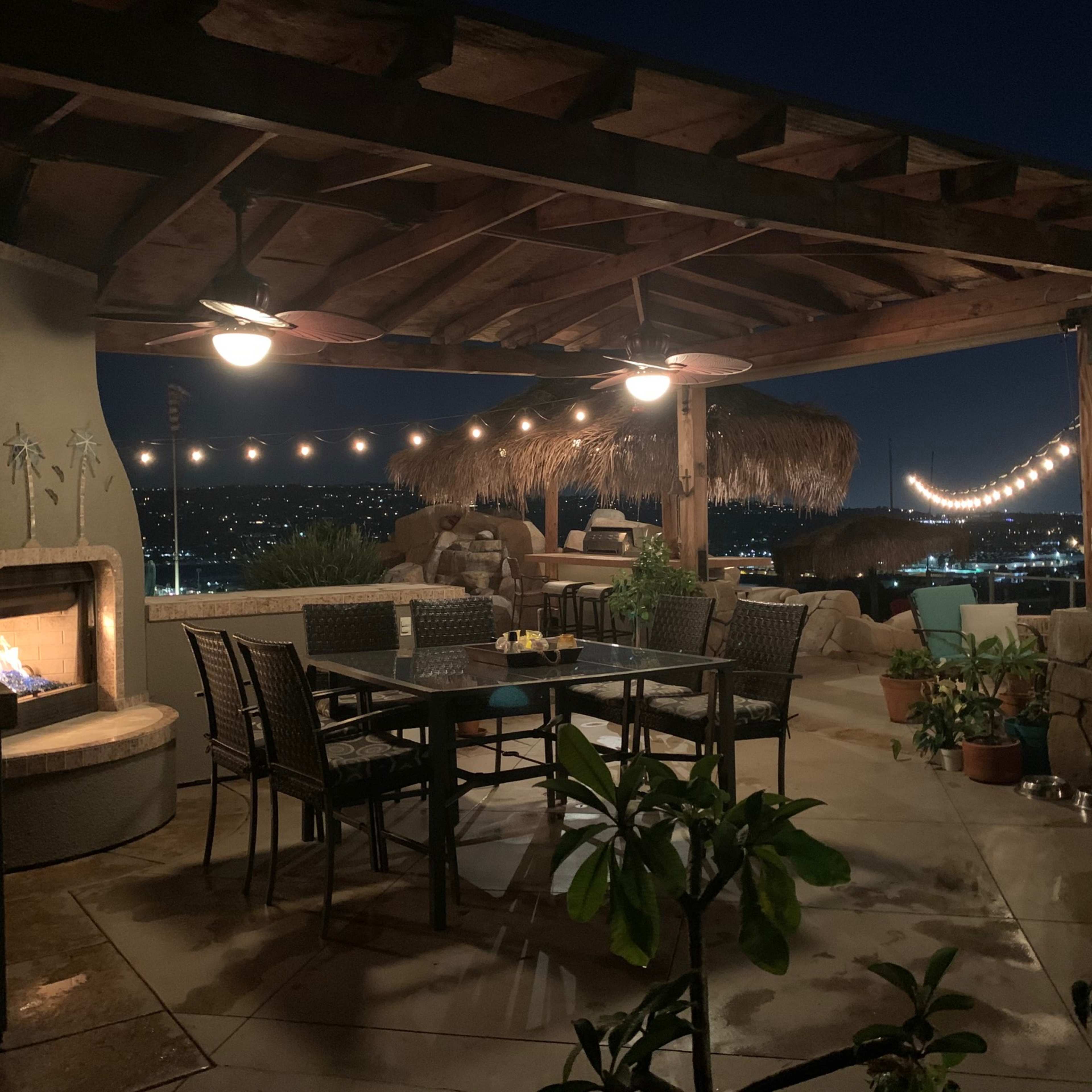 A patio at night with a dining table surrounded by chairs, a fire feature, and a thatched-roof lounge area, overlooking a city skyline adorned with lights.