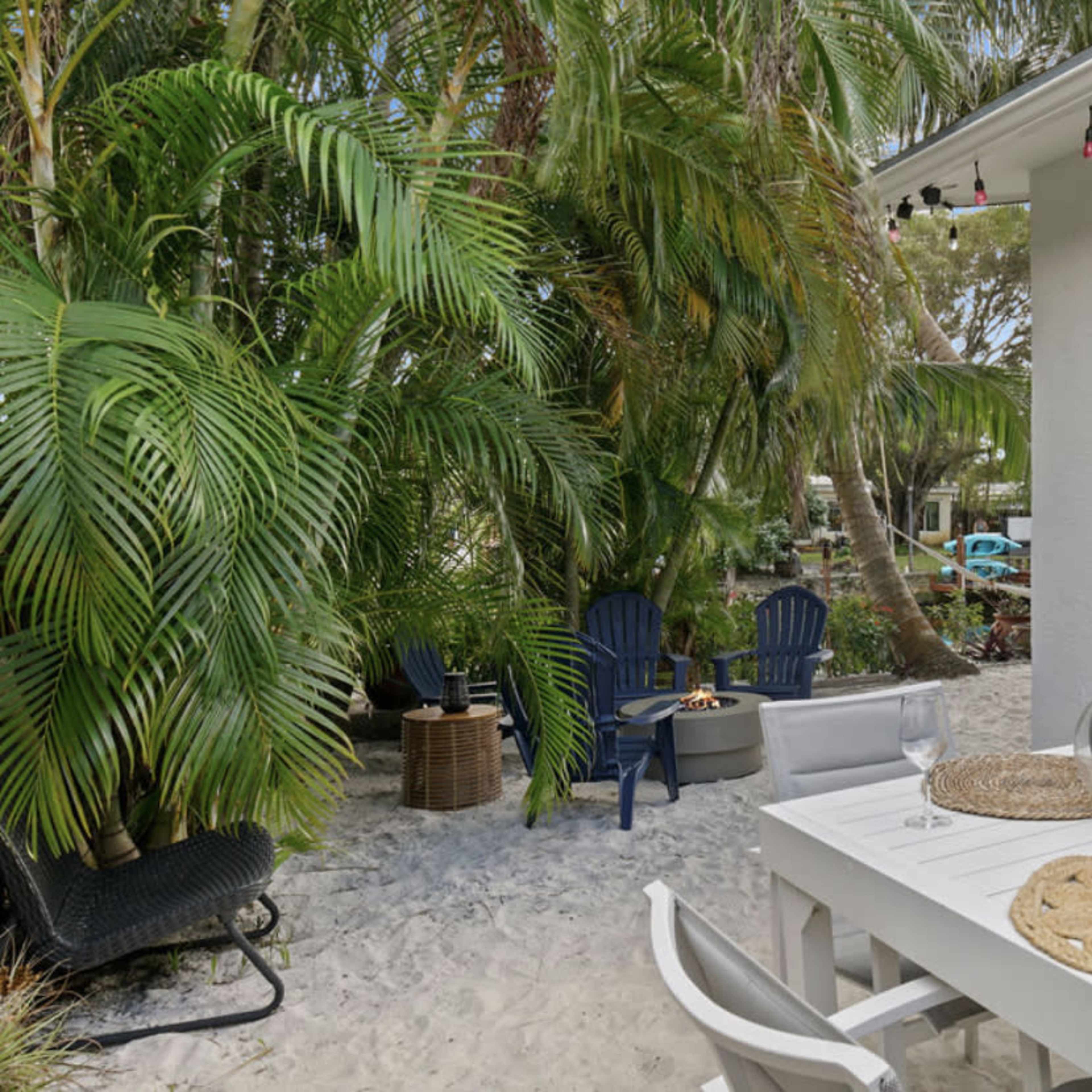 A white outdoor dining table is set with glasses and a bottle of wine near a sandy area surrounded by palm trees and lounge chairs.