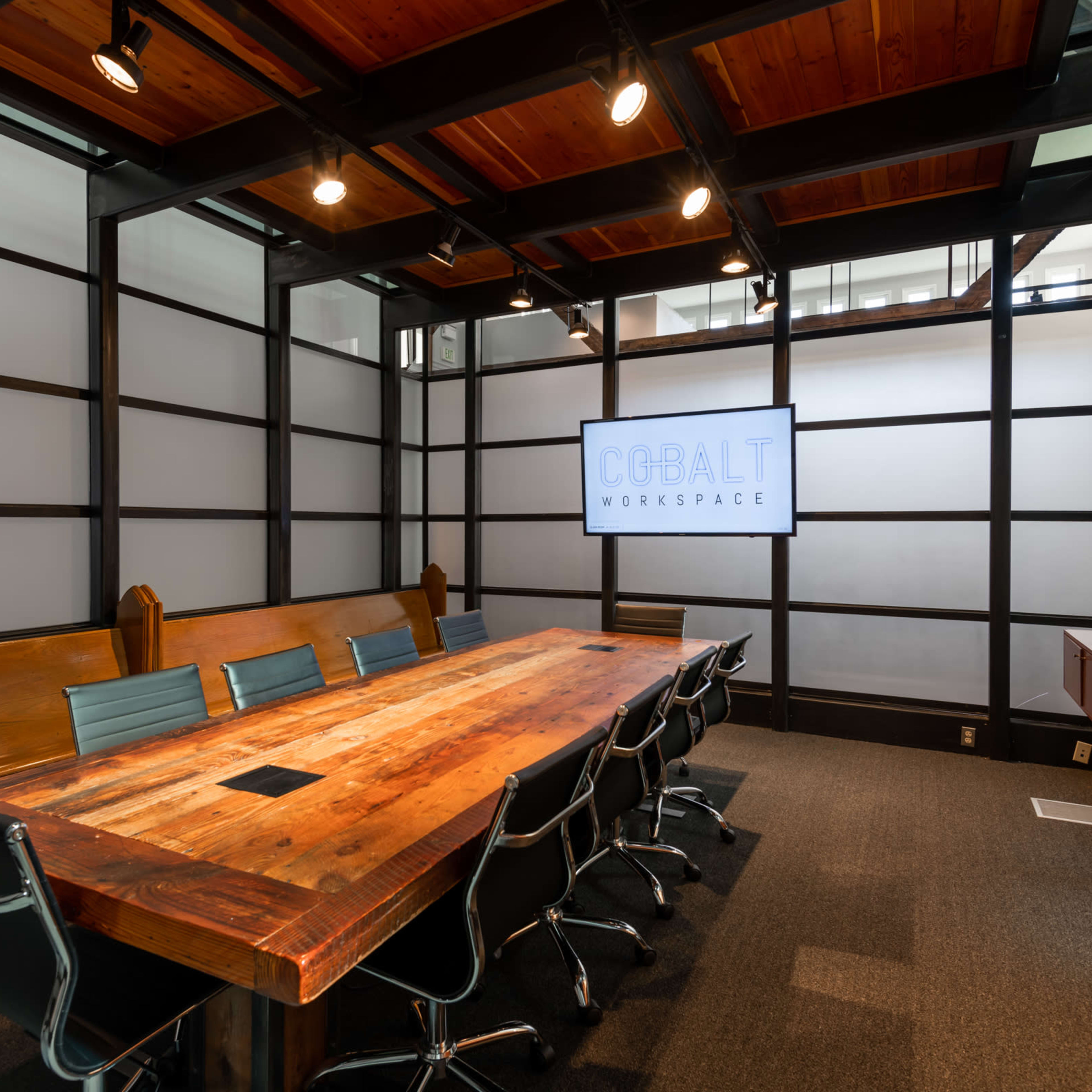 A modern conference room with a large wooden table, several black office chairs, and a wall-mounted display screen.