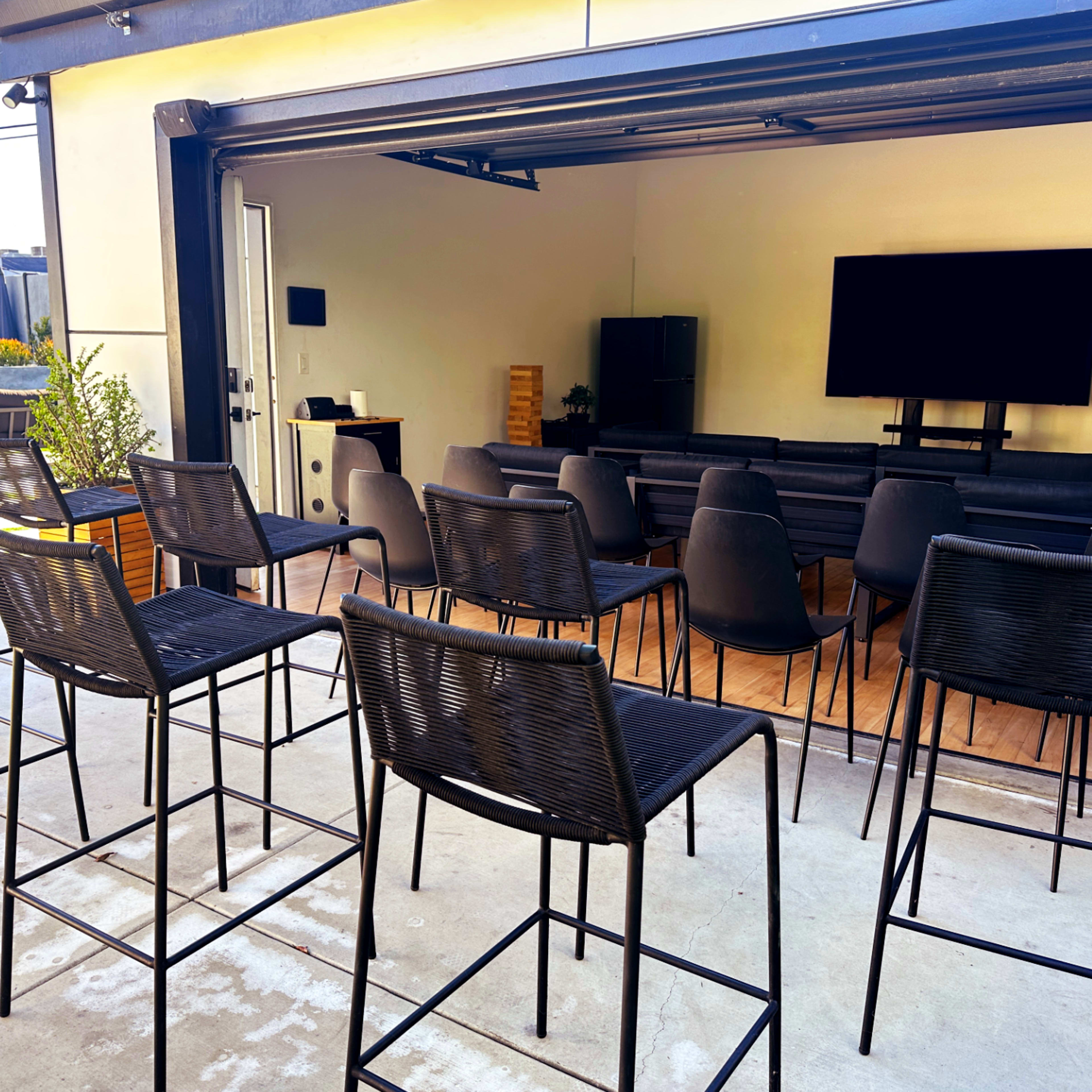A modern outdoor seating area with high-top chairs facing a large screen, alongside views of a pool and additional lounge areas.