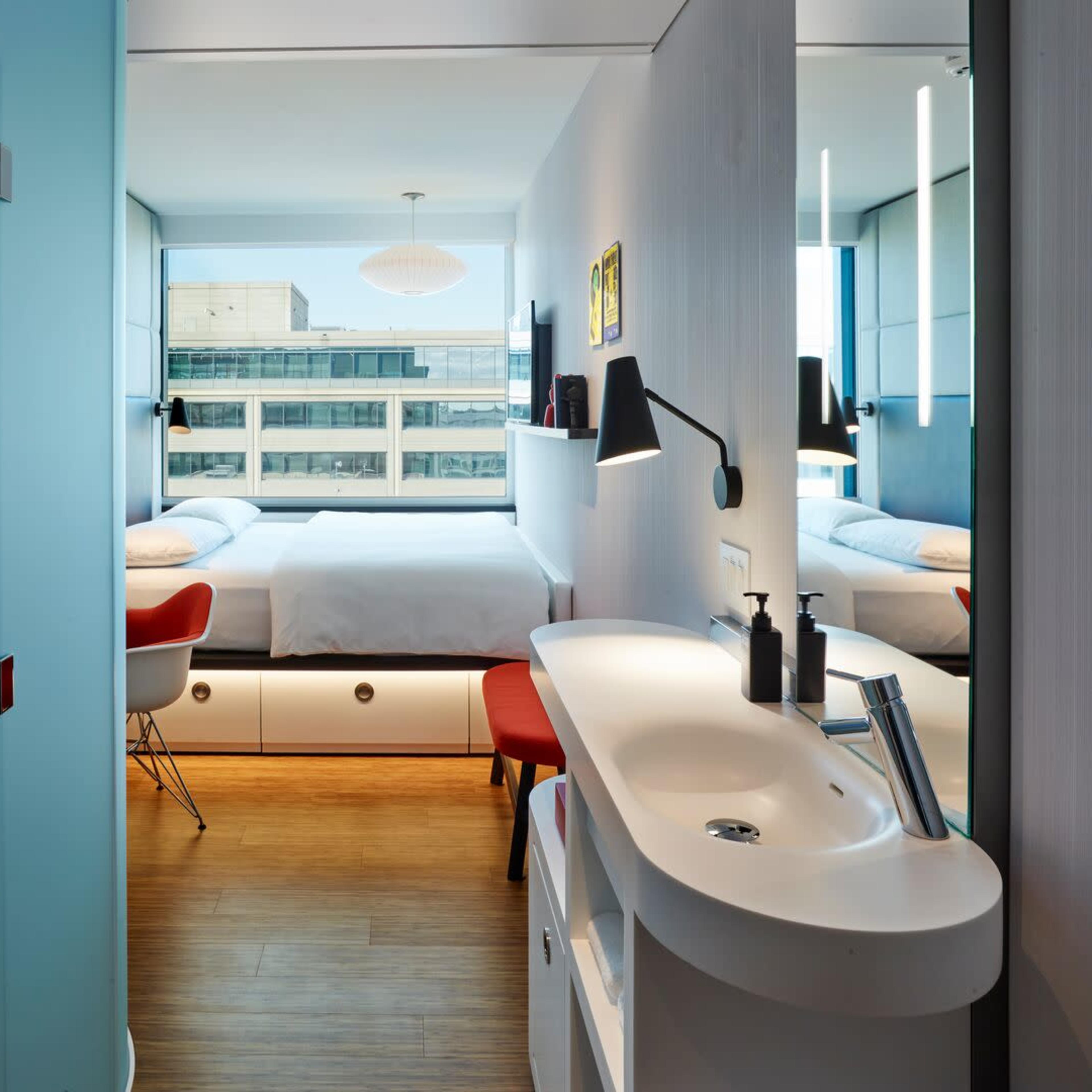 A modern hotel room with a bed, a sink area, and large windows offering a view of the building outside.