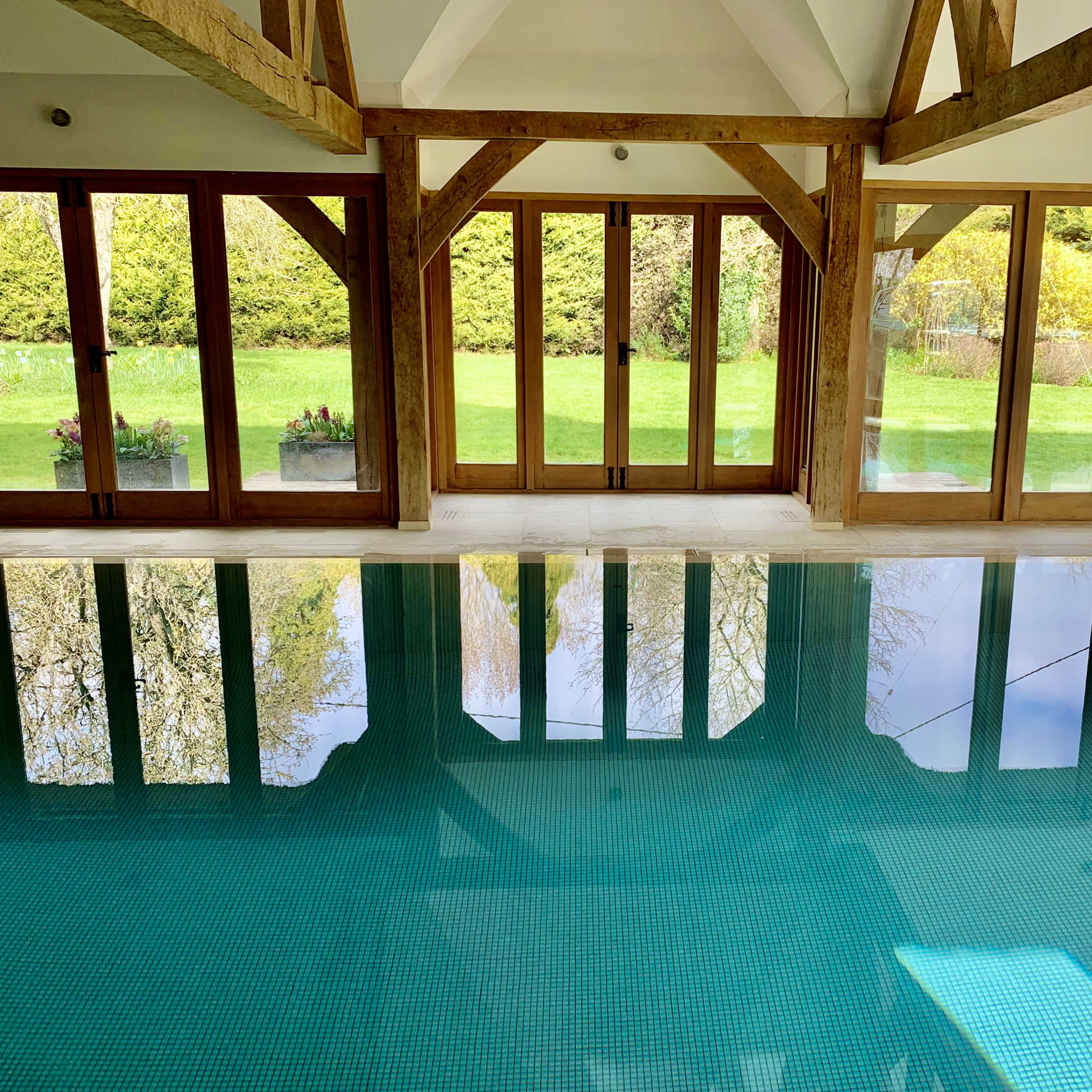 A modern indoor pool area with wooden beams and large glass doors leading to a grassy outdoor space.