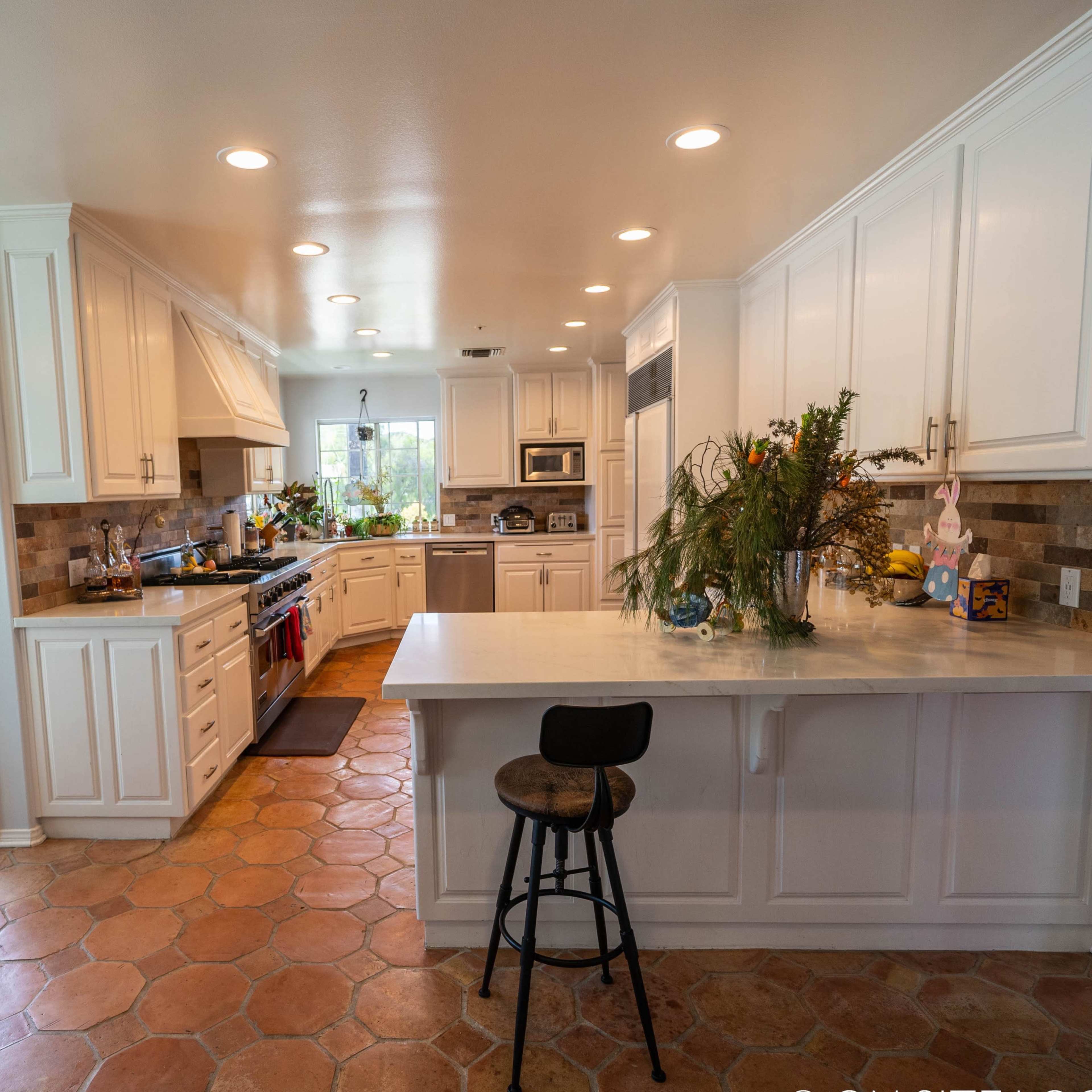 The kitchen features white cabinetry, a spacious island with a stool, and terracotta tile flooring.