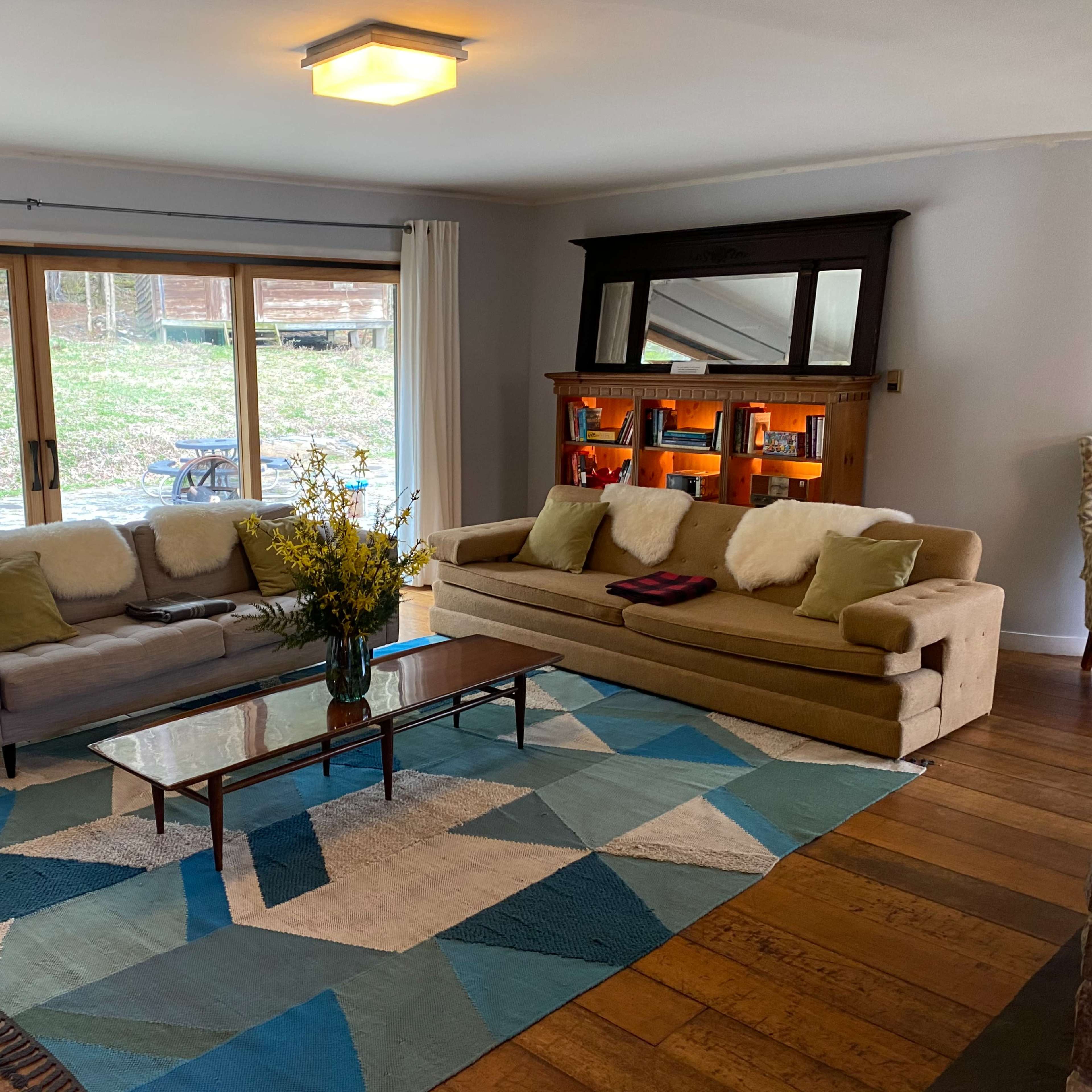 A living room with two sofas facing a coffee table, a patterned area rug on the wooden floor, and large sliding glass doors that open to a garden area.