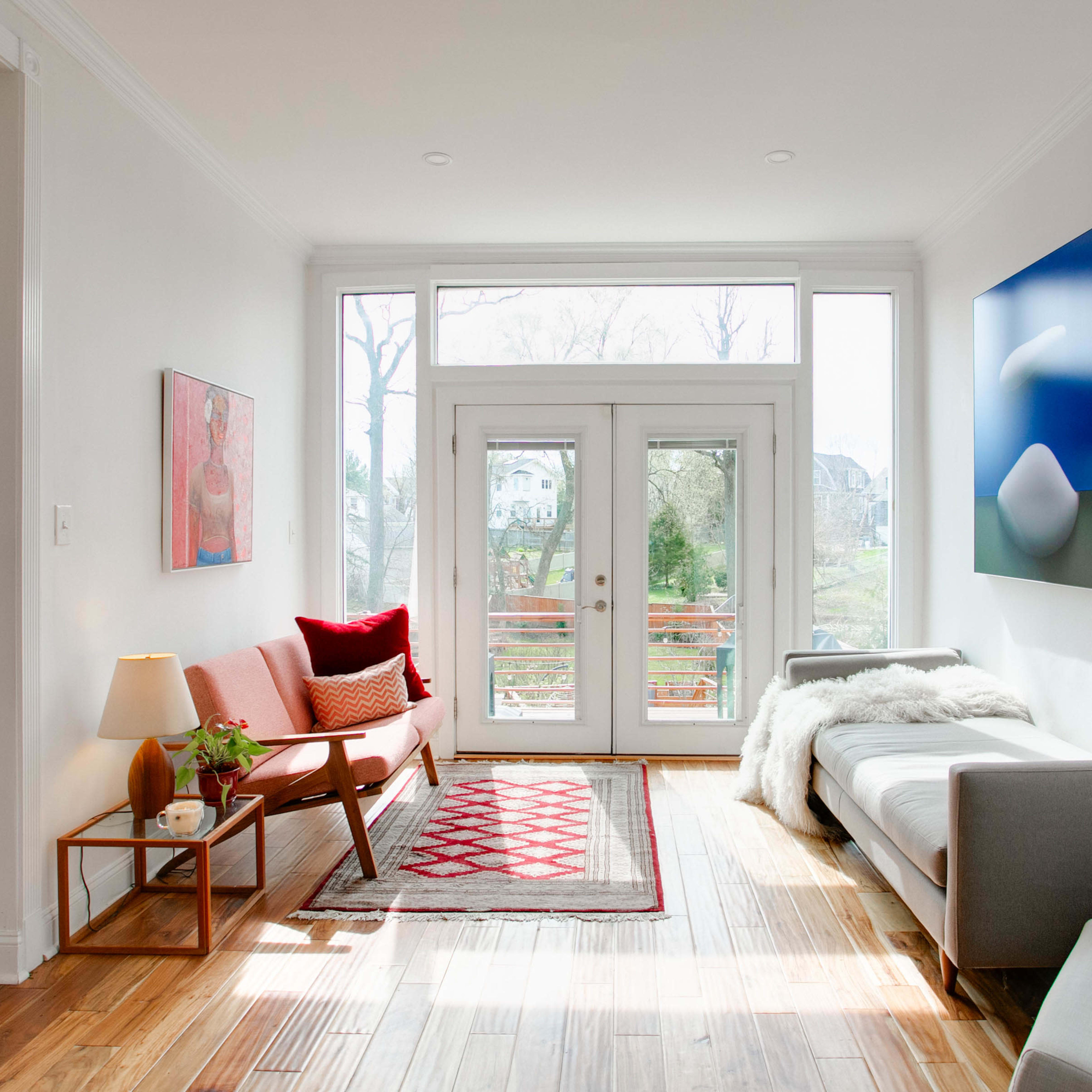 A bright living room features a pink sofa, a gray couch, a coffee table, and large windows overlooking a garden.