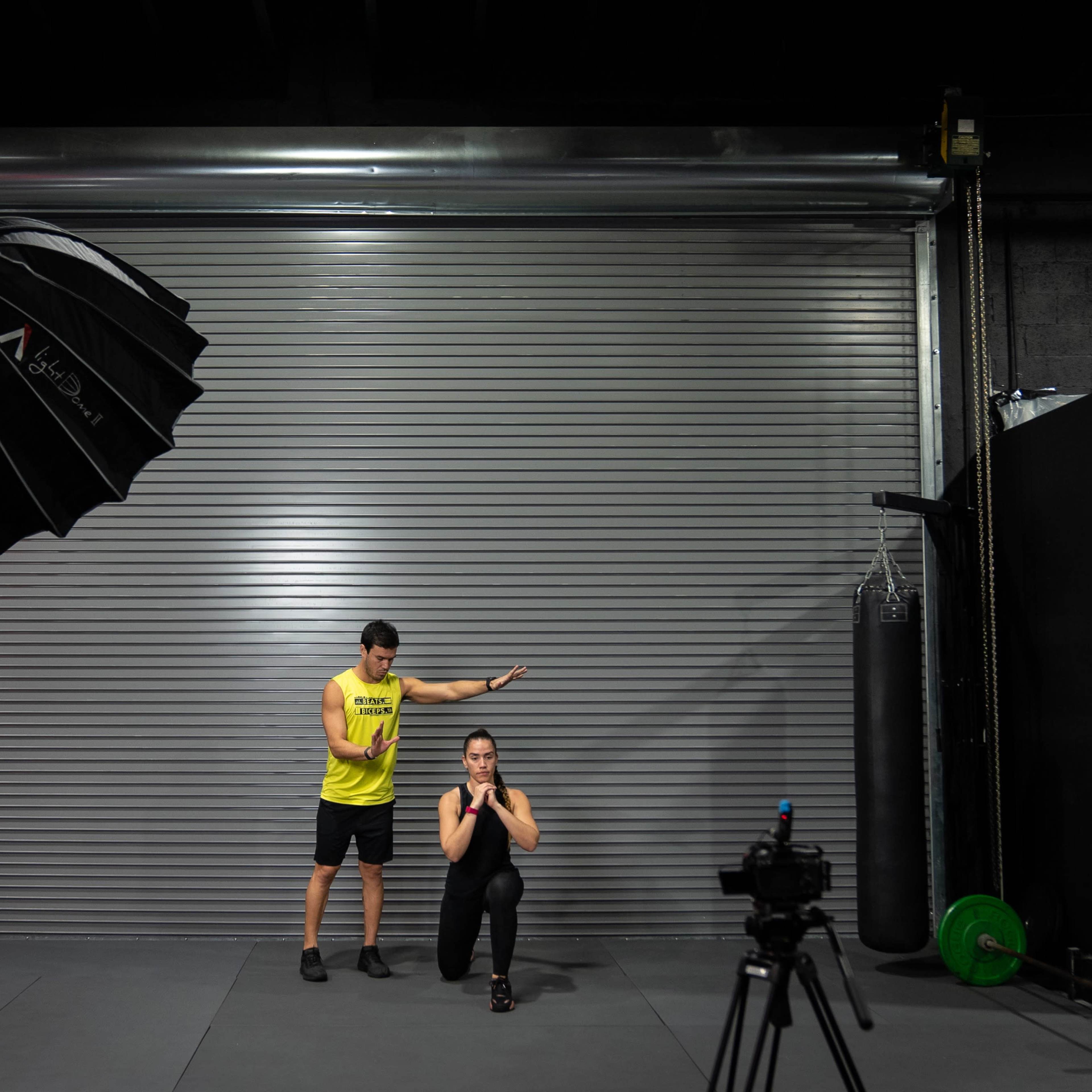 A man in a yellow tank top stands beside a woman in a squat position in a gym setting with fitness equipment visible.