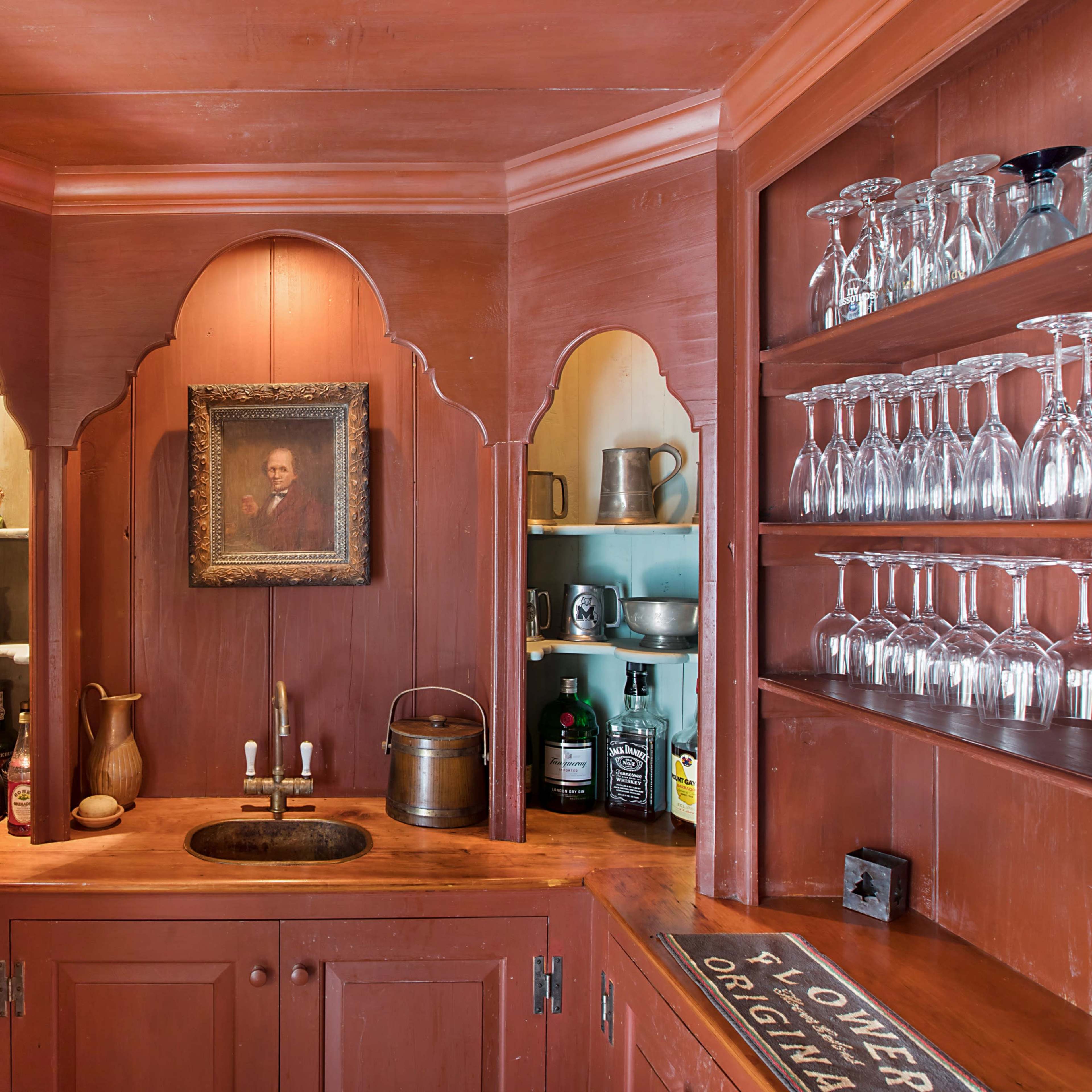 A wooden bar area featuring a sink, shelves lined with glasses and bottles, and a framed portrait on the wall.