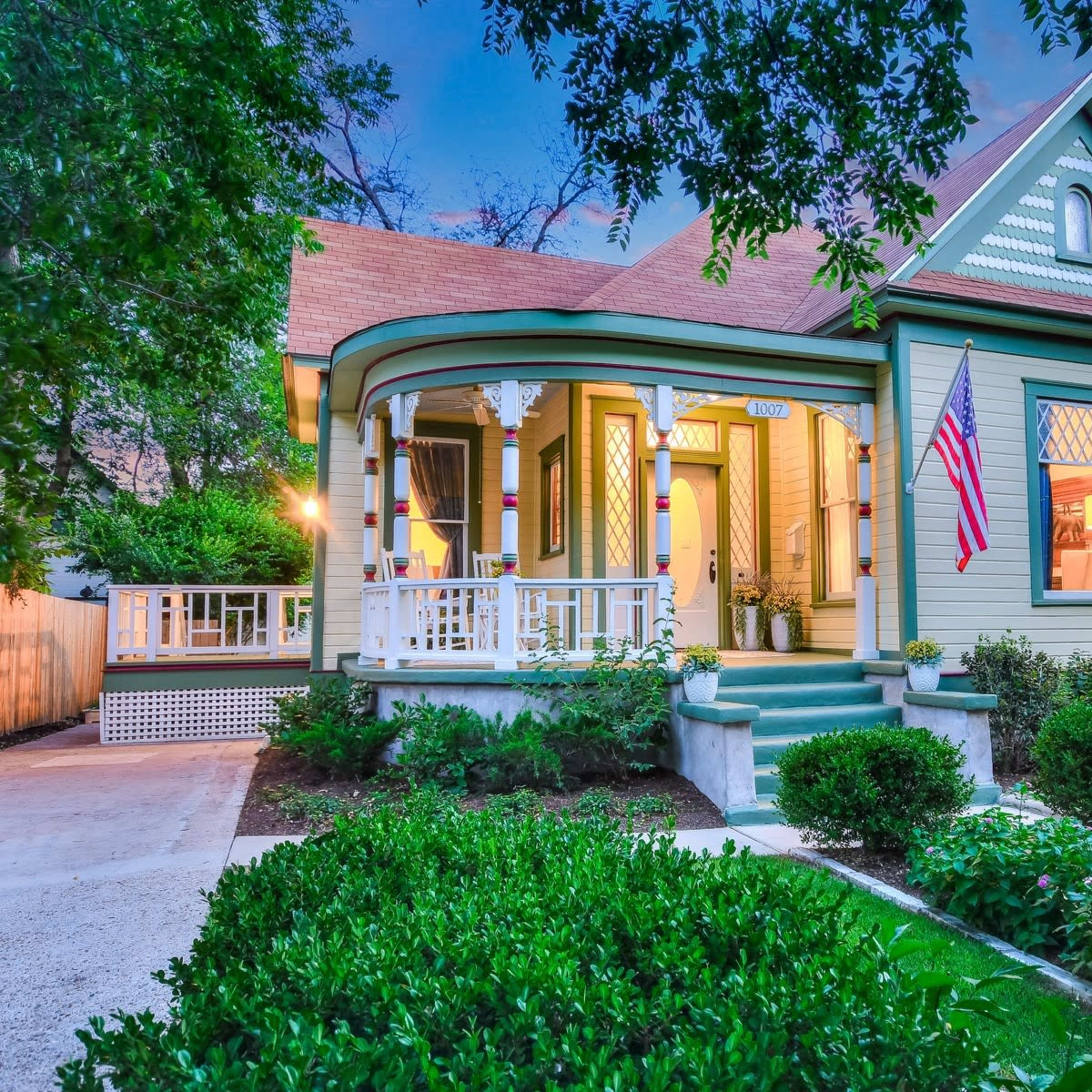 A Victorian-style house with an inviting porch, surrounded by well-maintained gardens and trees, features an American flag displayed prominently.