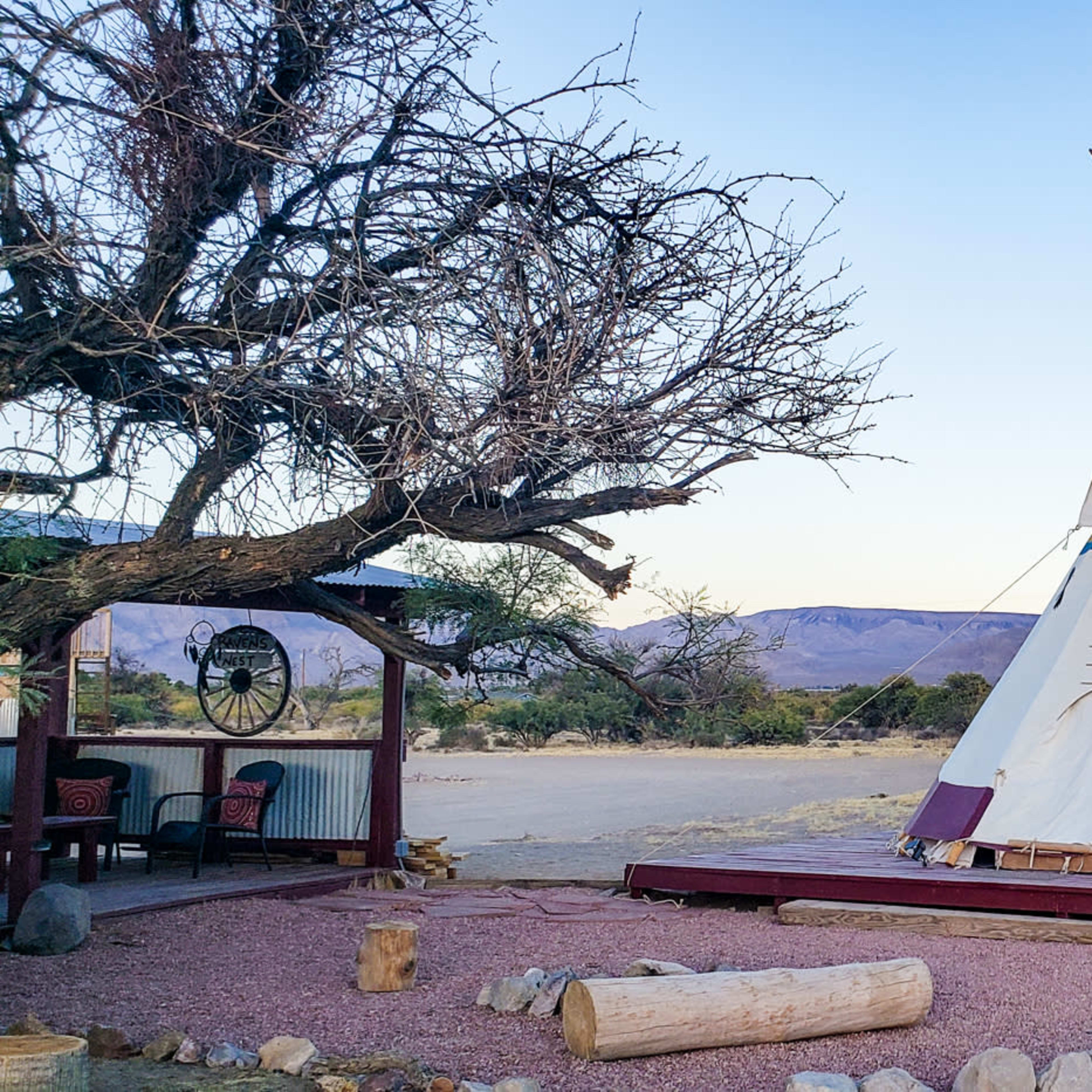 A teepee stands on a wooden platform surrounded by shrubs and distant mountains under a clear sky.