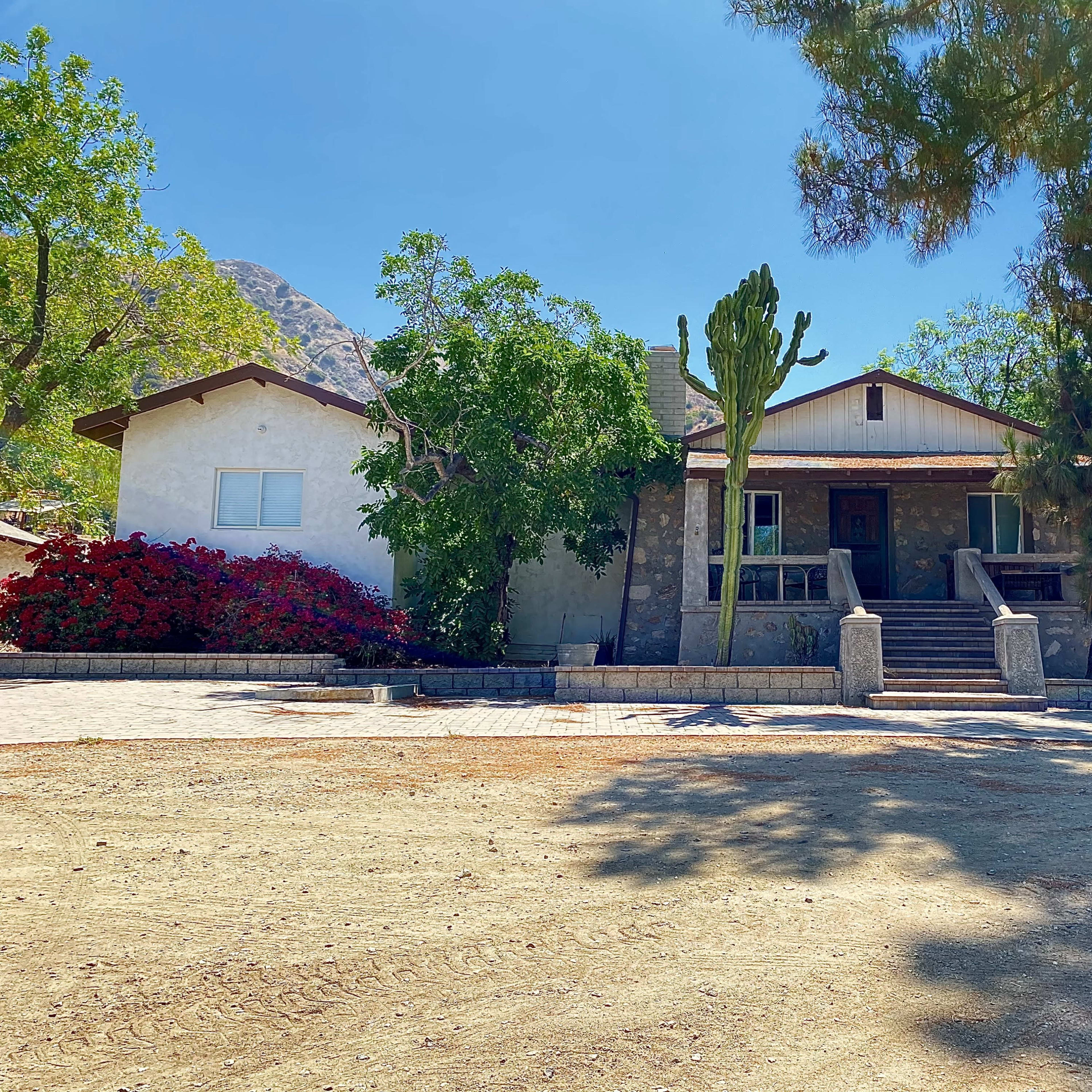A two-story house is set against a backdrop of mountains, surrounded by trees and flowering plants, with a gravel driveway leading to the entrance.