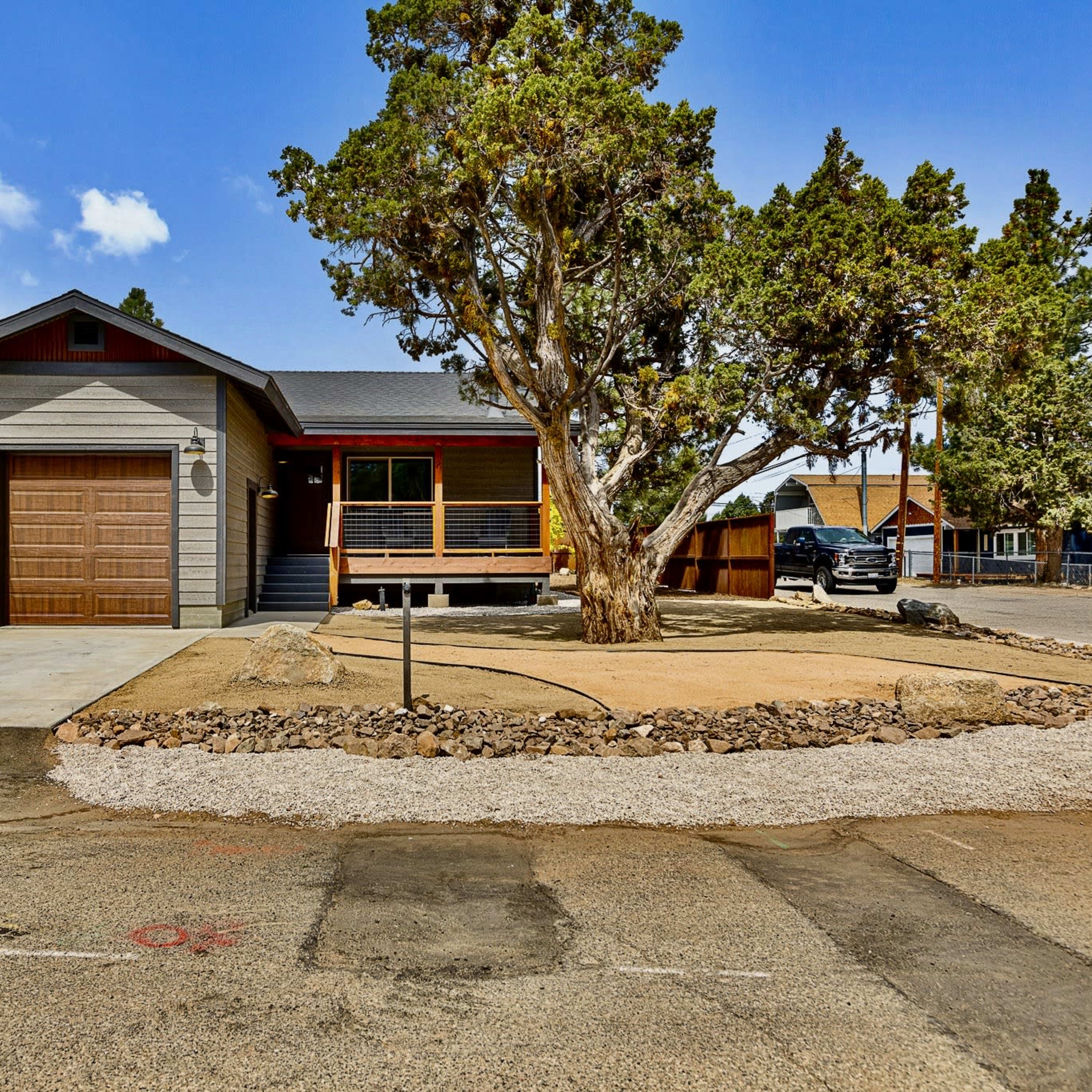A single-story house with a wooden garage door and a covered porch is surrounded by trees and a gravel landscaping area.