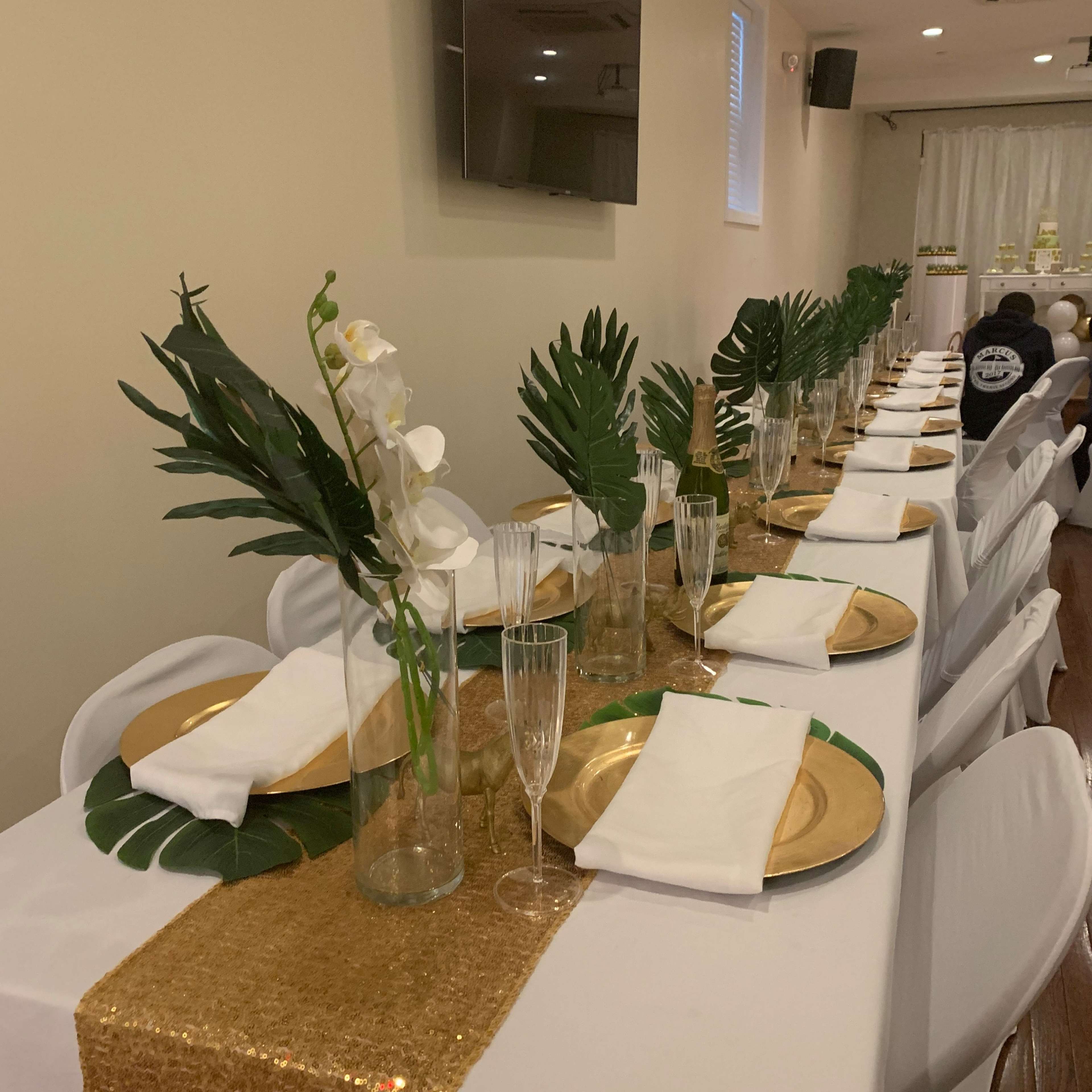 A long dining table is set with gold plates, champagne flutes, white napkins, and decorative greenery under a gold sequined table runner.