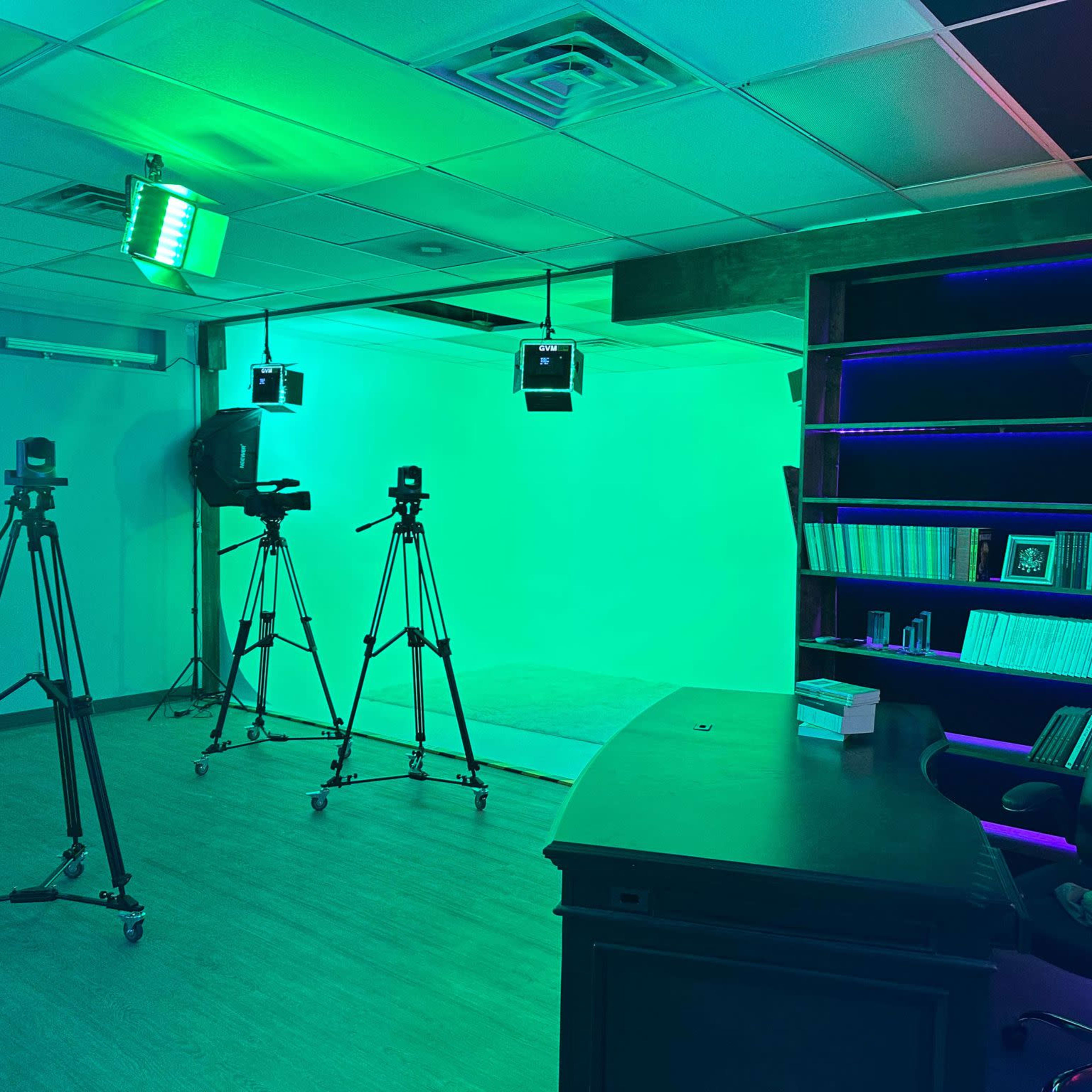 A studio setup with three tripods for cameras, a green backdrop, and a desk with an ergonomic chair.