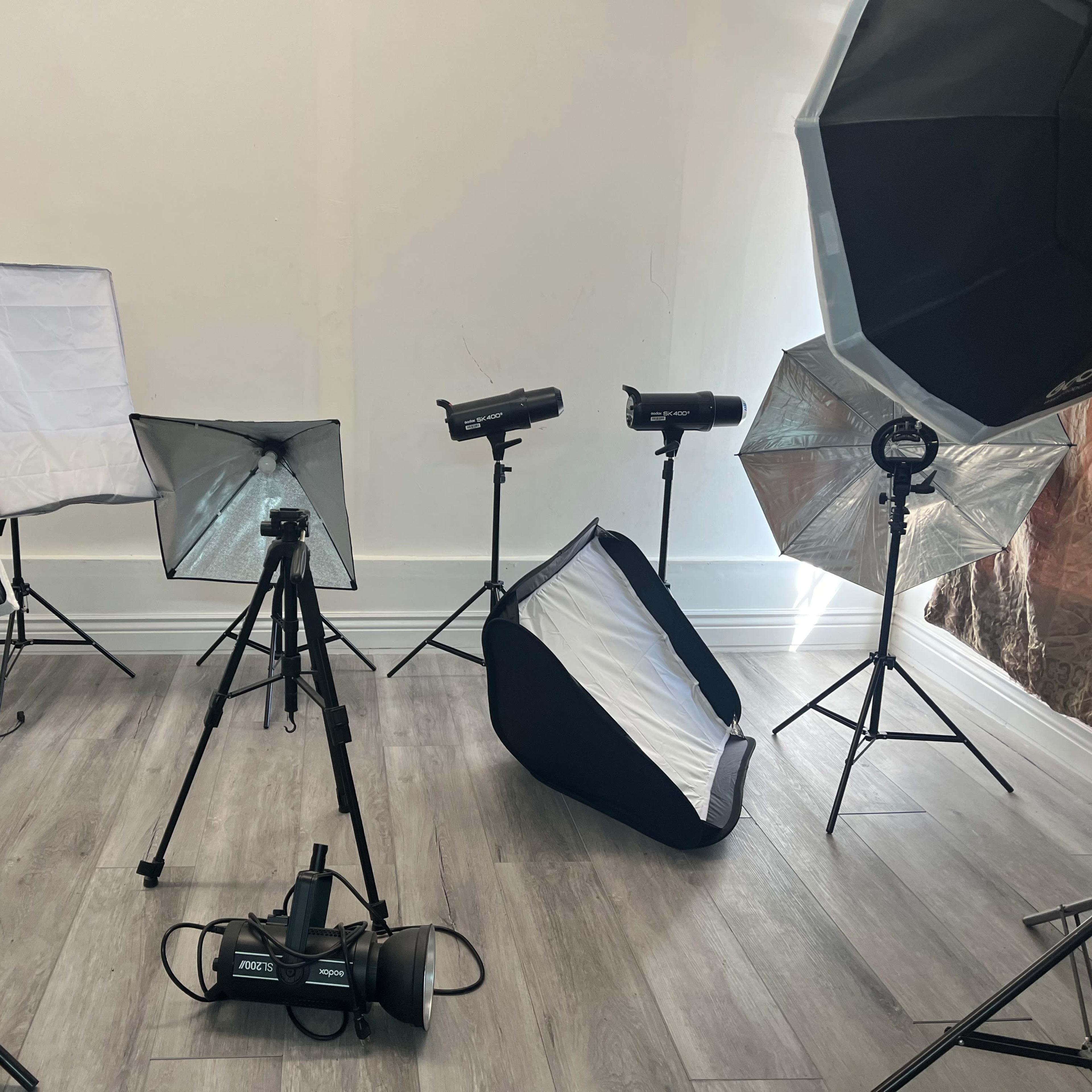 A photography studio setup with various lighting equipment, softboxes, and a tripod arranged around a light-colored backdrop.