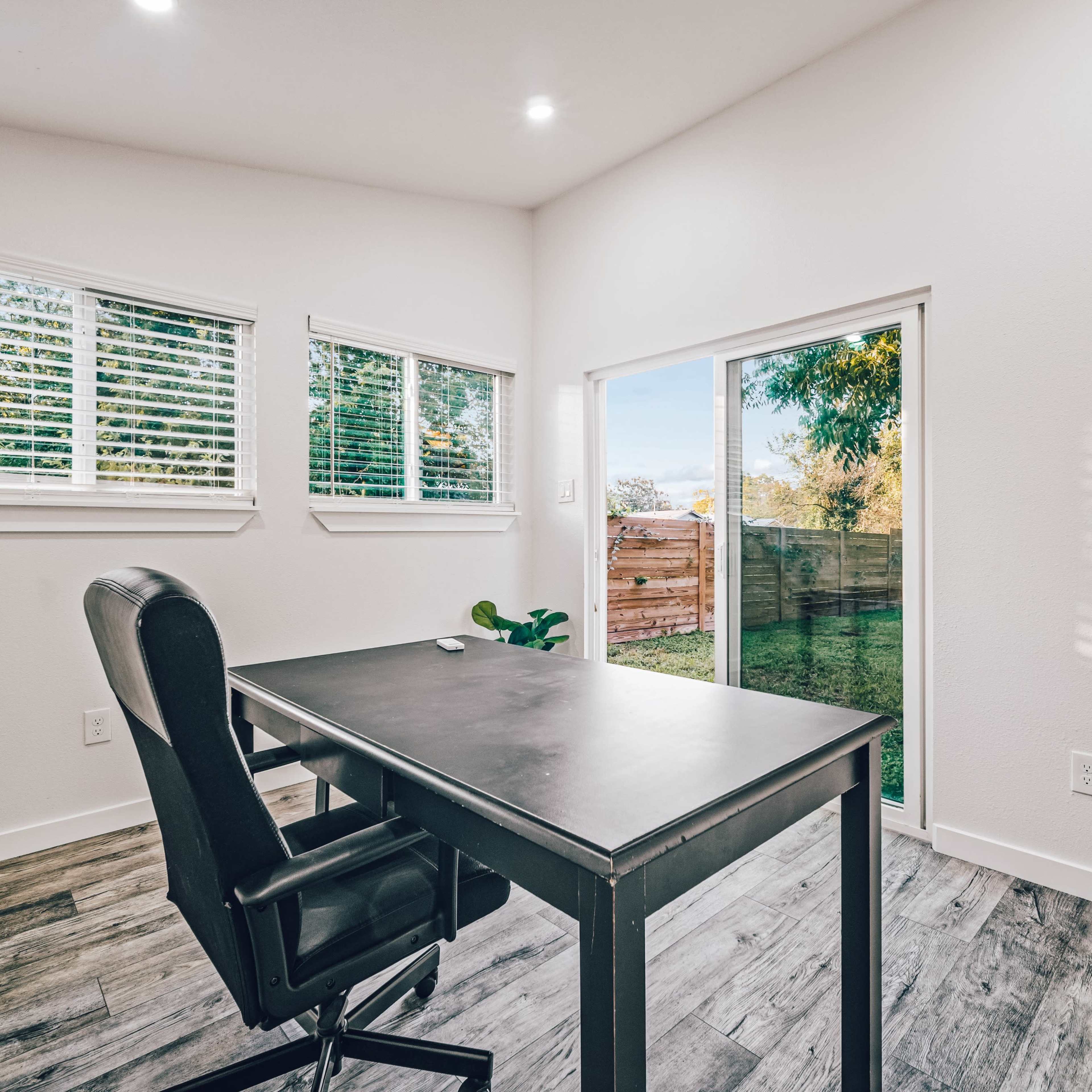 A modern office space features a black desk and chair, with large windows allowing natural light and a view of a fenced outdoor area.