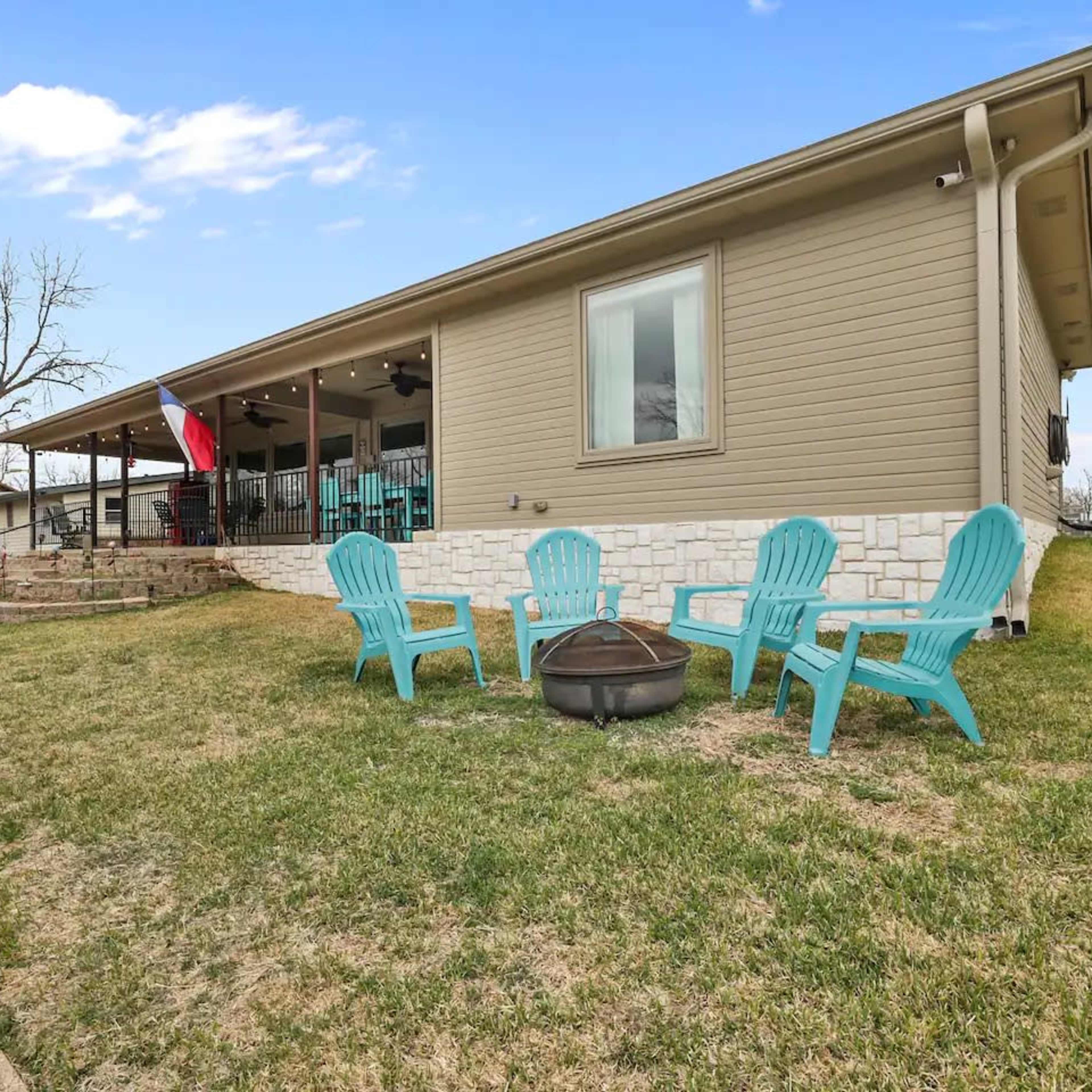 A single-story house with a stone foundation features a patio area with four blue chairs and a fire pit in the grassy yard.