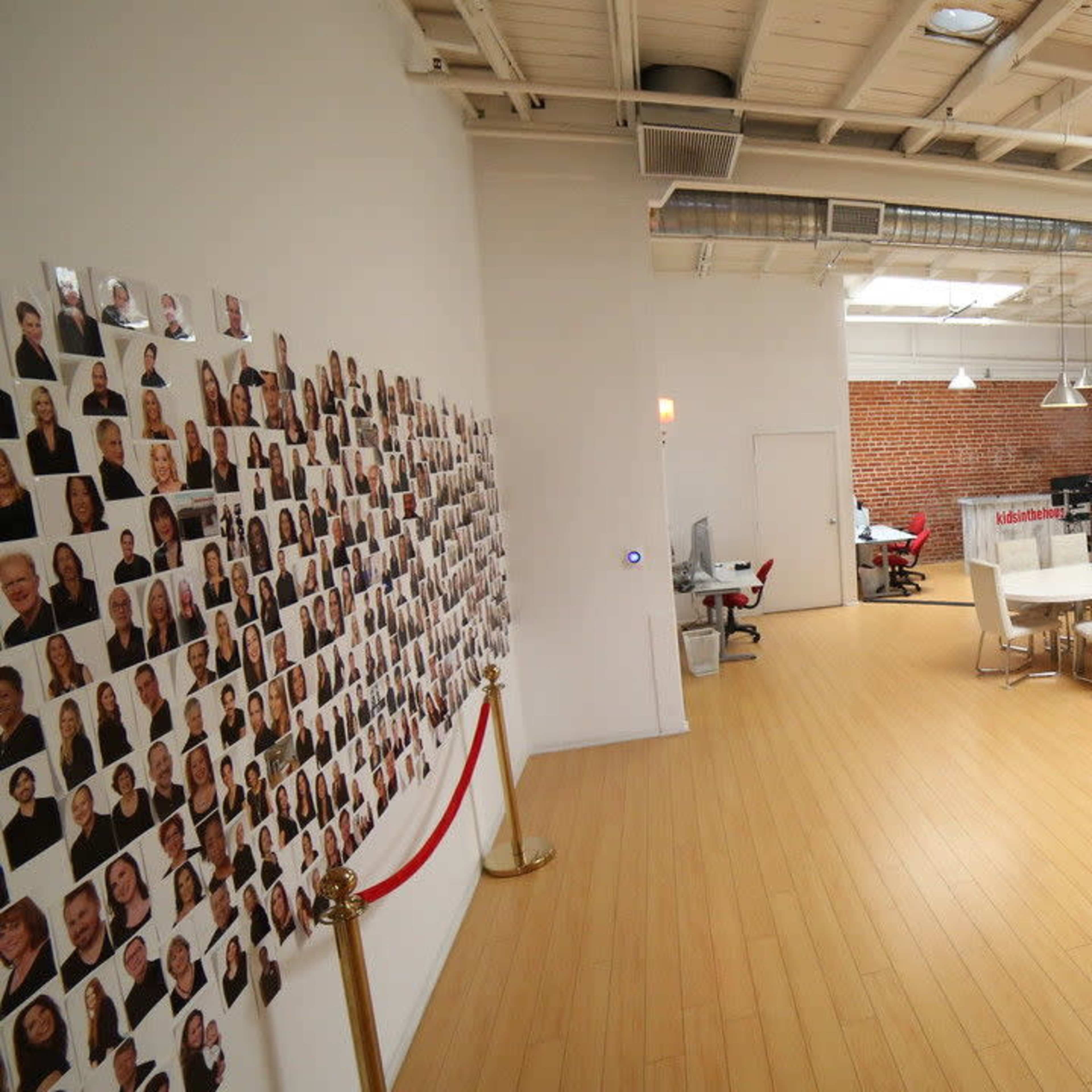A large modern office space with a polished wooden floor, featuring a long table surrounded by white chairs, a wall covered with numerous photographs, and exposed brick walls.